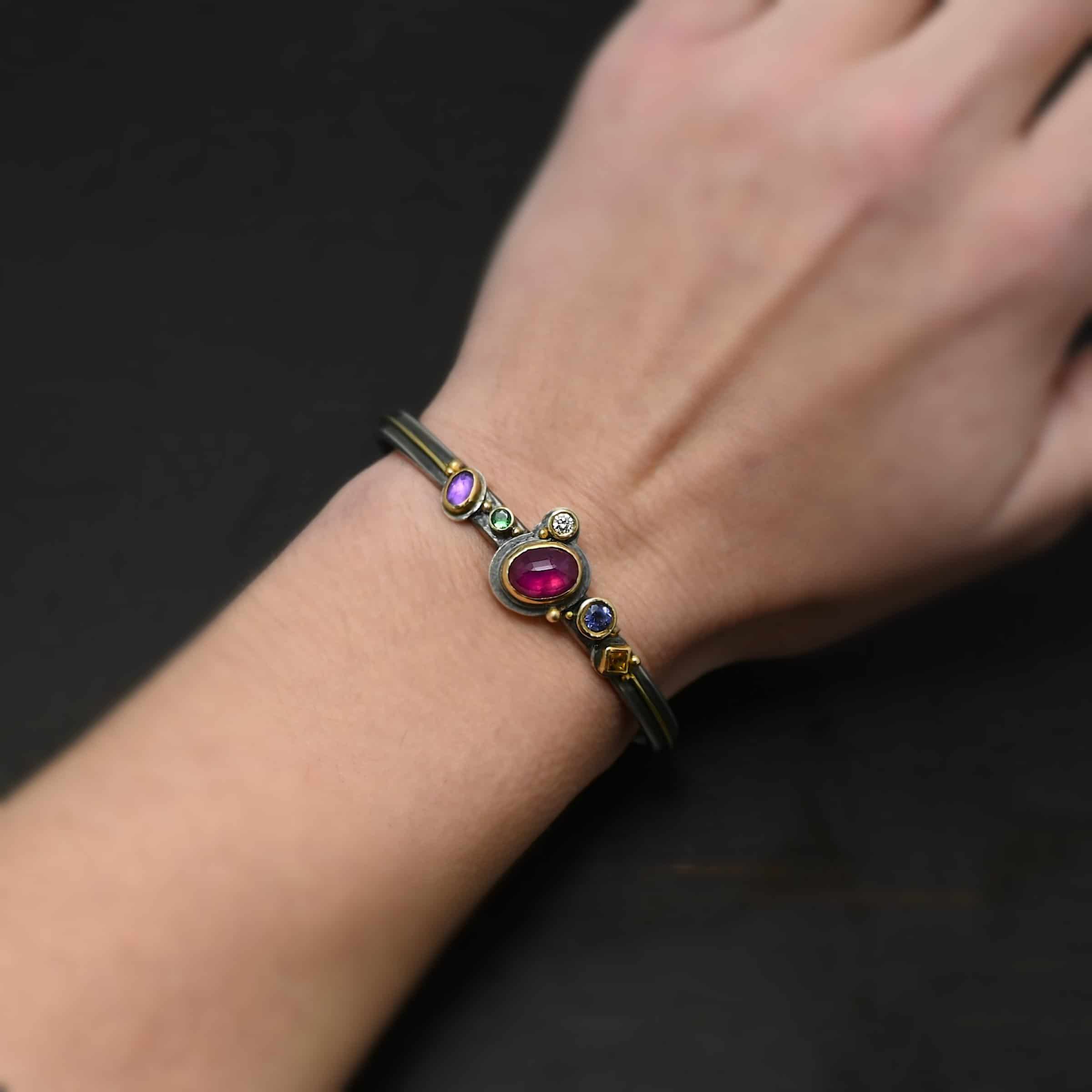 Cinderella Cuff Photo of the "Cinderella" Cuff. It has a ruby in the center, oval shaped and set sideways. There is an amethyst and tsavorite garnet on the left side and a citrine and amethyst on the right side. There is a diamond on the top left side of the ruby. Each stone is set in gold. There is a thin line of gold going across the cuff through the center on each side where the stones aren't. There are balls of gold scattered around the stones. Cuff is worn by a model.