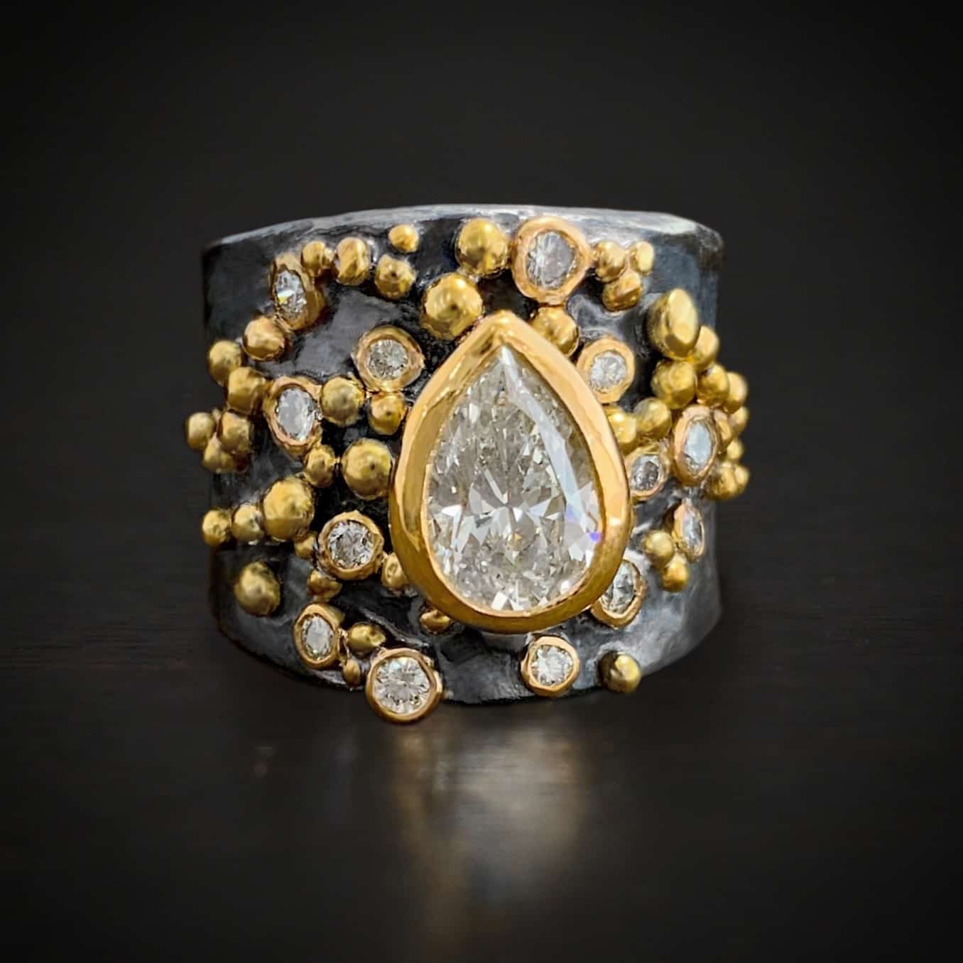 Photo of the "Starstruck" Engagement Ring. There is a pear shaped moissanite in the center of the ring, point facing up. It is set in gold. There are multiple diamonds scattered around the ring, all set in gold. There are multiple balls of gold around the entire ring, put closely together.