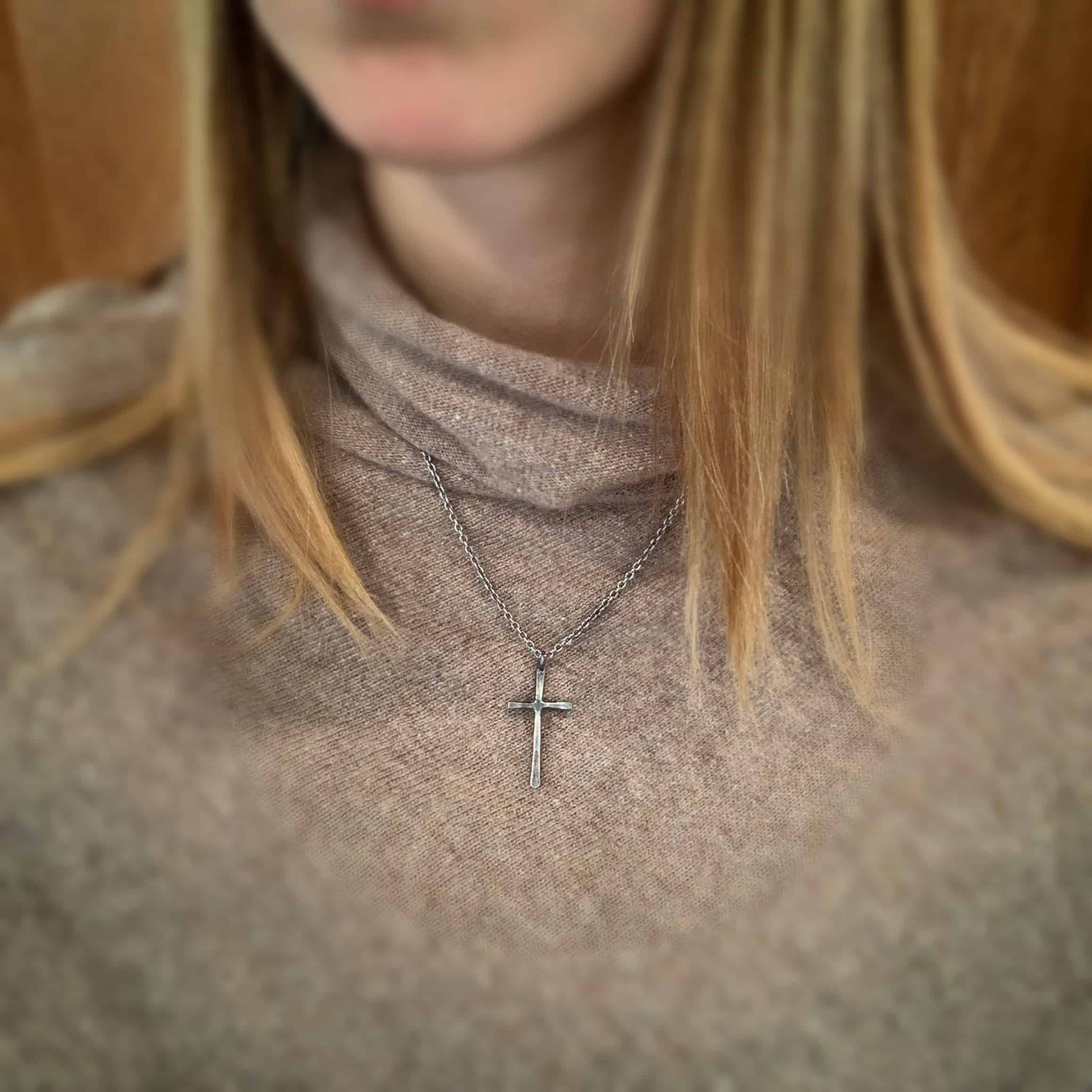 Photo of the "Healer" Necklace. Made out of sterling silver. There is a medium sized cross that is hammered flat, but smooth. It is oxidized and has a loop soldered to the top of it. There is a chain that goes through the top of the necklace. Necklace is worn by a model.
