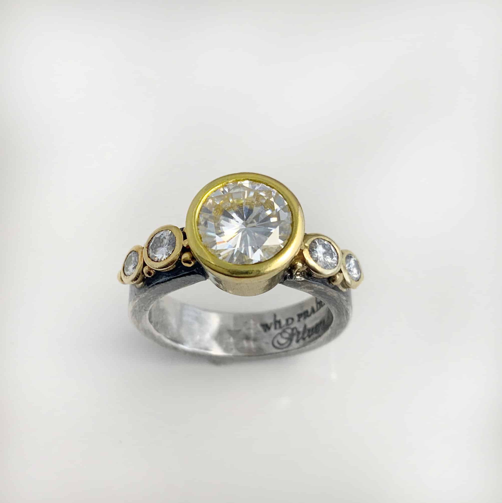 Until Forever Ring Round 4 Photo of the "Until Forever" Engagement Ring. There is a large moissanite in the center of the ring. There are four more moissanites that are smaller, two on each side of the center moissanite. There are balls of gold scattered around each moissanite.