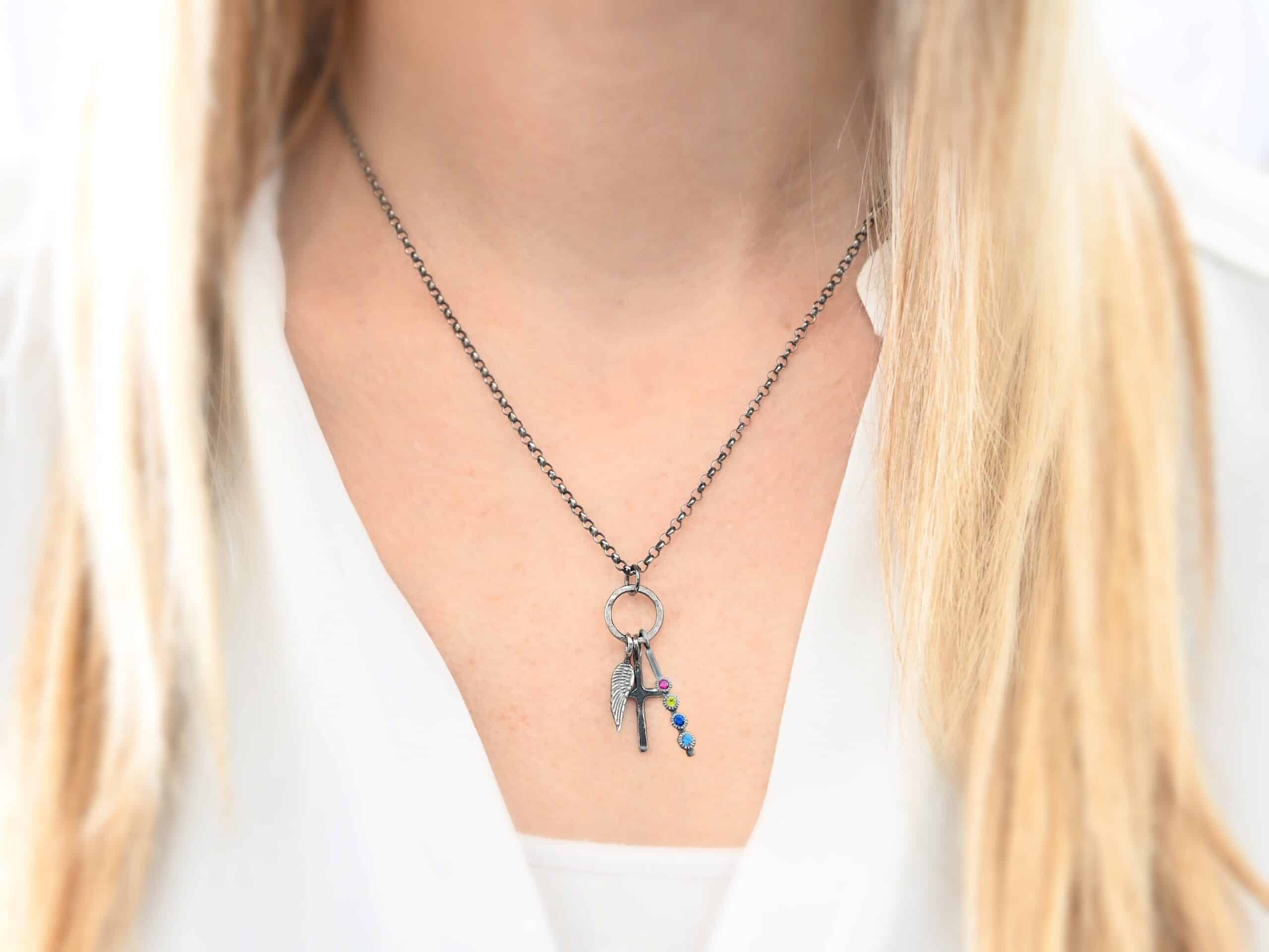 Photo of a cross and angel wing necklace. Made out of oxidized sterling silver. There is a small sized cross that is flat and smooth. It has a loop soldered to the top of it. There is a small sized angel wing pendant next to it, also with a soldered loop on the top. There is a small, long rectangle with four birthstones on it and a soldered loop on the top. The birthstones are ruby, peridot, sapphire, and aquamarine. There is a chain that goes through the top of the necklace that is silver and oxidized. Necklace is worn by a model.
