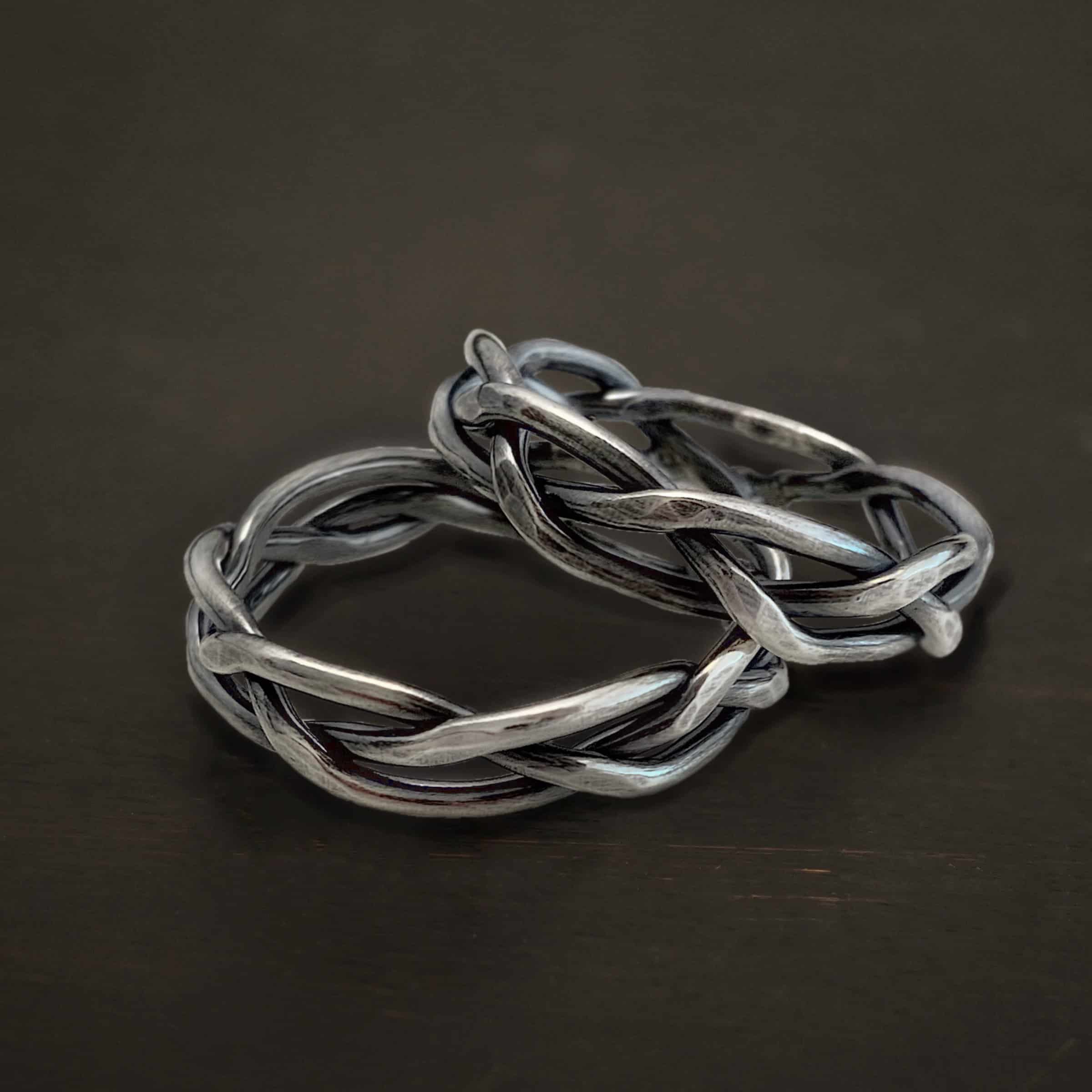 Bekah Braided Band Photo of the "Braided Bekah" Band. Bands are made out of sterling silver. The bands are made out of three lines that are hammered. They all weave and intertwine with one another to create the band. There are two bands, one resting on top of the other. Bands are sitting against a dark background.