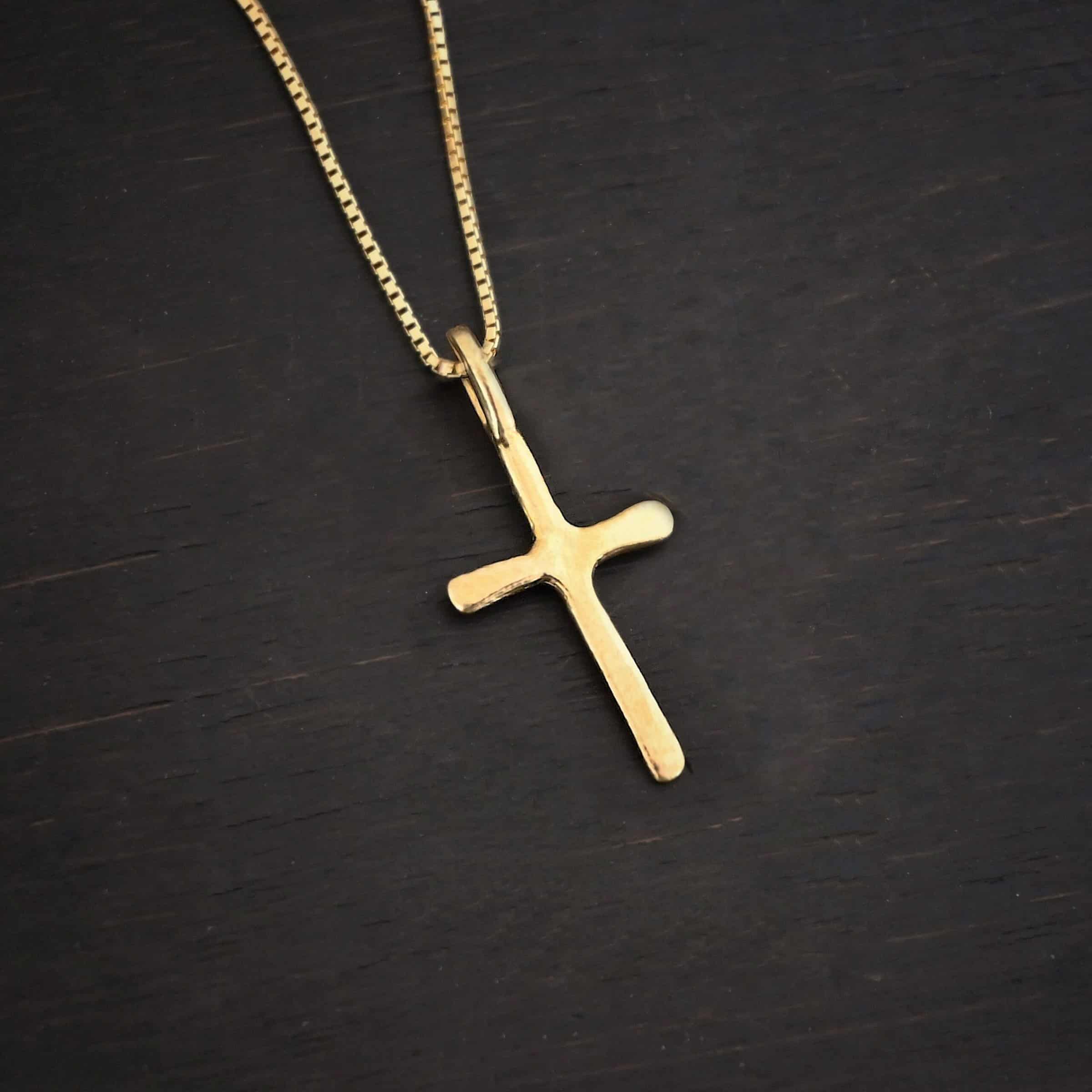 Divine Necklace Photo of the "Divine" Necklace. Made out of 18k gold. There is a small sized cross that is flat and smooth. It has a loop soldered to the top of it. There is a chain that goes through the loop of the cross. The chain is also gold. Necklace is lying against a dark background.