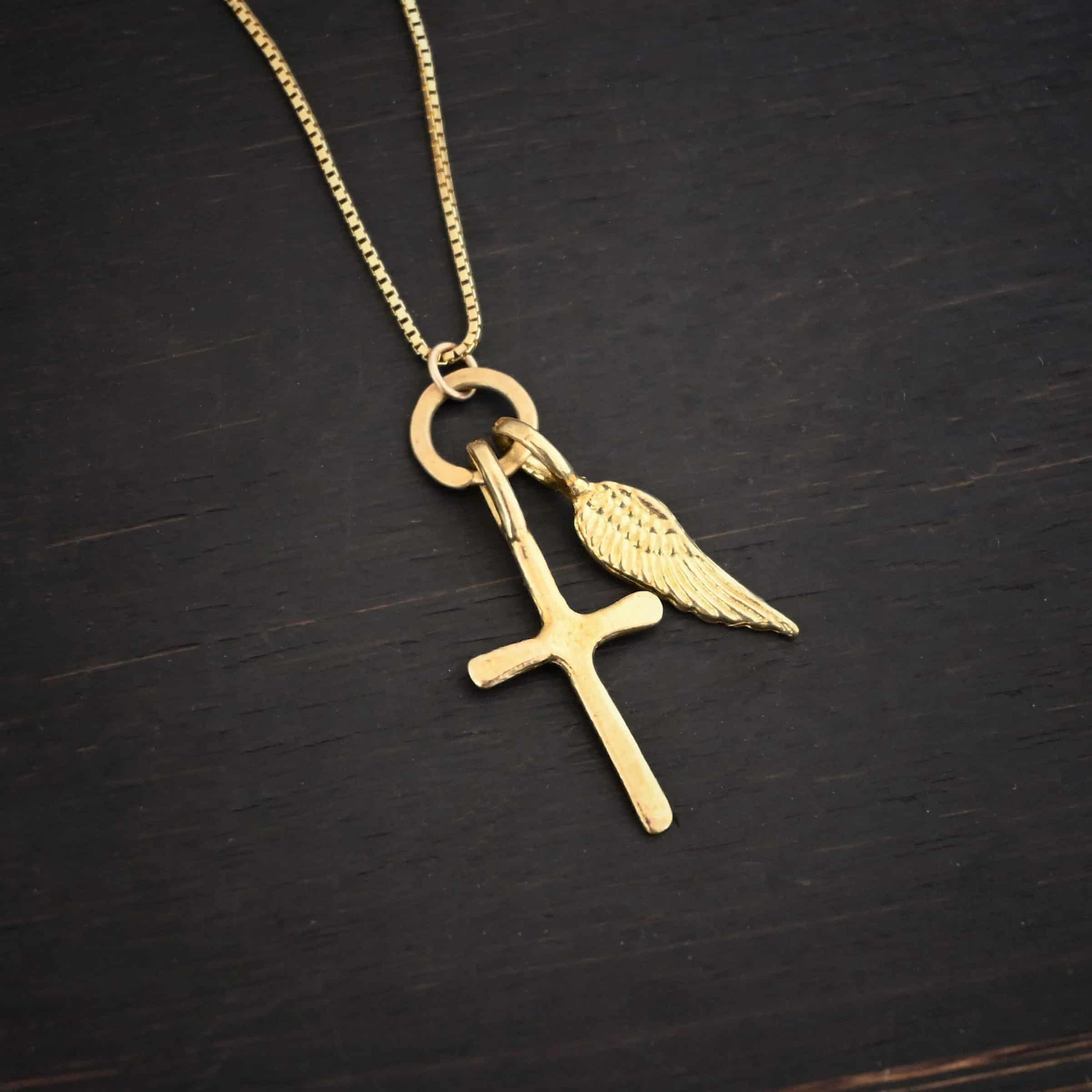 Divine Necklace Photo of the "Divine" Necklace. Made out of 18k gold. There is a small sized cross that is flat and smooth. It has a loop soldered to the top of it. There is a small sized angel wing pendant next to it, also with a soldered loop on the top. There is a chain that goes through the top of the necklace that is also gold. Necklace is lying against a dark background.