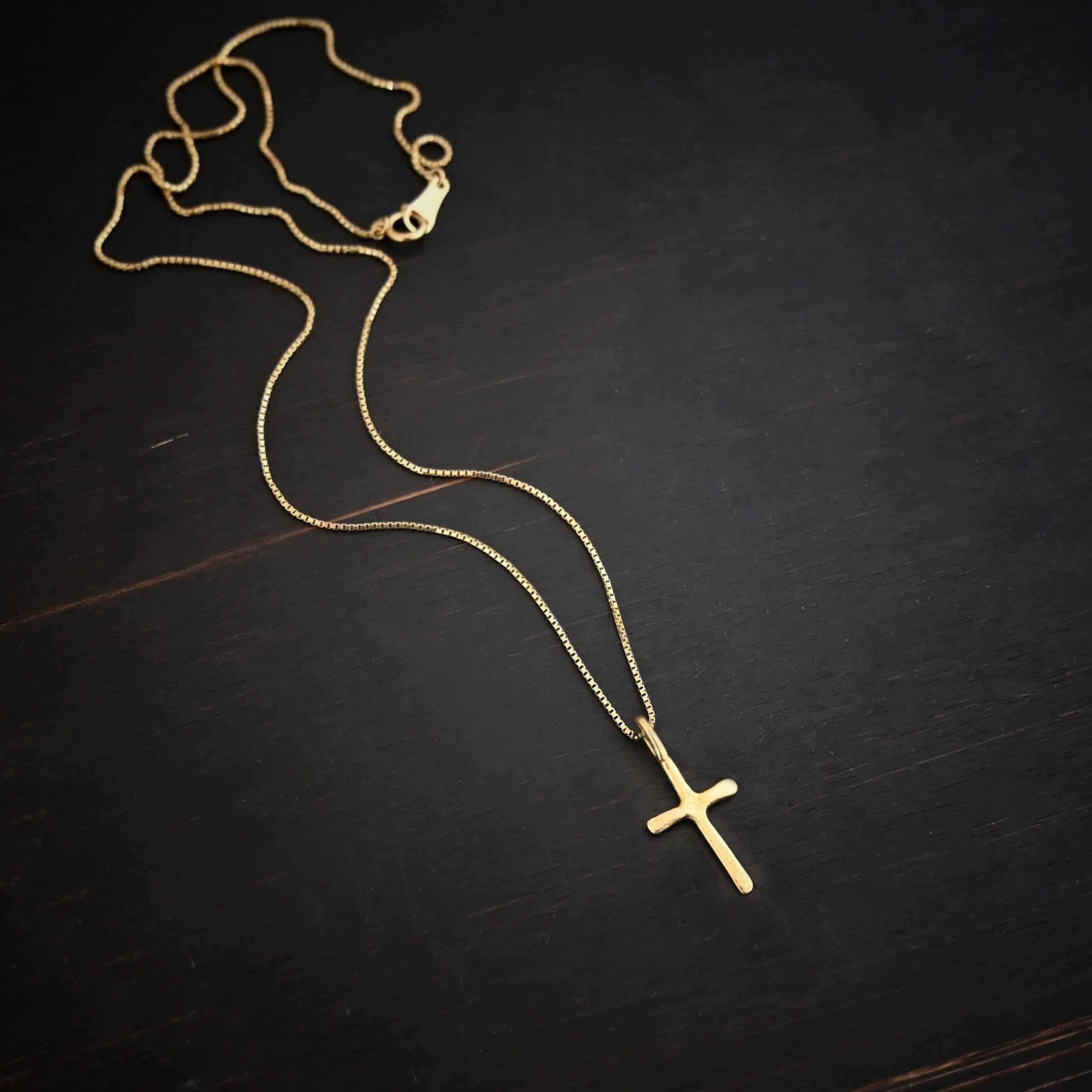 Divine Necklace Photo of the "Divine" Necklace. Made out of 18k gold. There is a small sized cross that is flat and smooth. It has a loop soldered to the top of it. There is a chain that goes through the loop of the cross. The chain is also gold. Necklace is lying against a dark background.