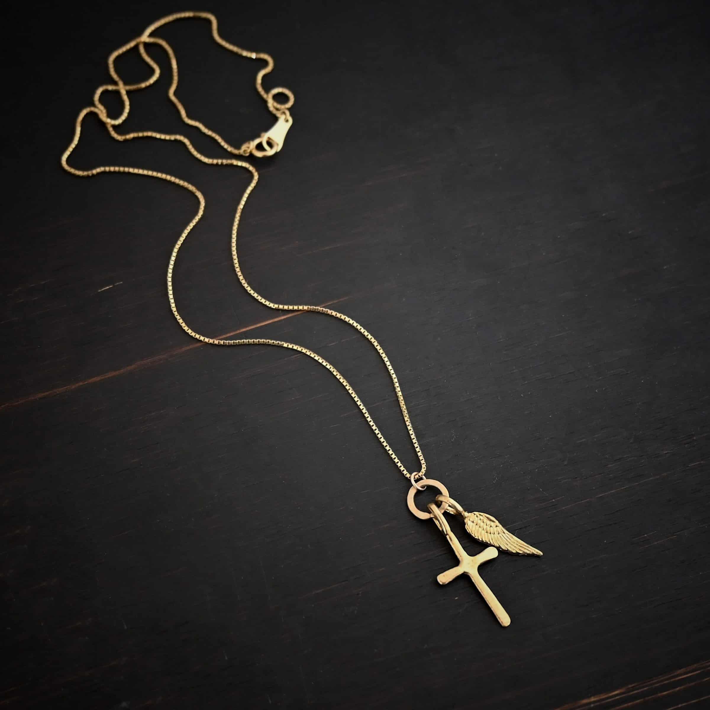 Divine Necklace Photo of the "Divine" Necklace. Made out of 18k gold. There is a small sized cross that is flat and smooth. It has a loop soldered to the top of it. There is a small sized angel wing pendant next to it, also with a soldered loop on the top. There is a chain that goes through the top of the necklace that is also gold. Necklace is lying against a dark background.