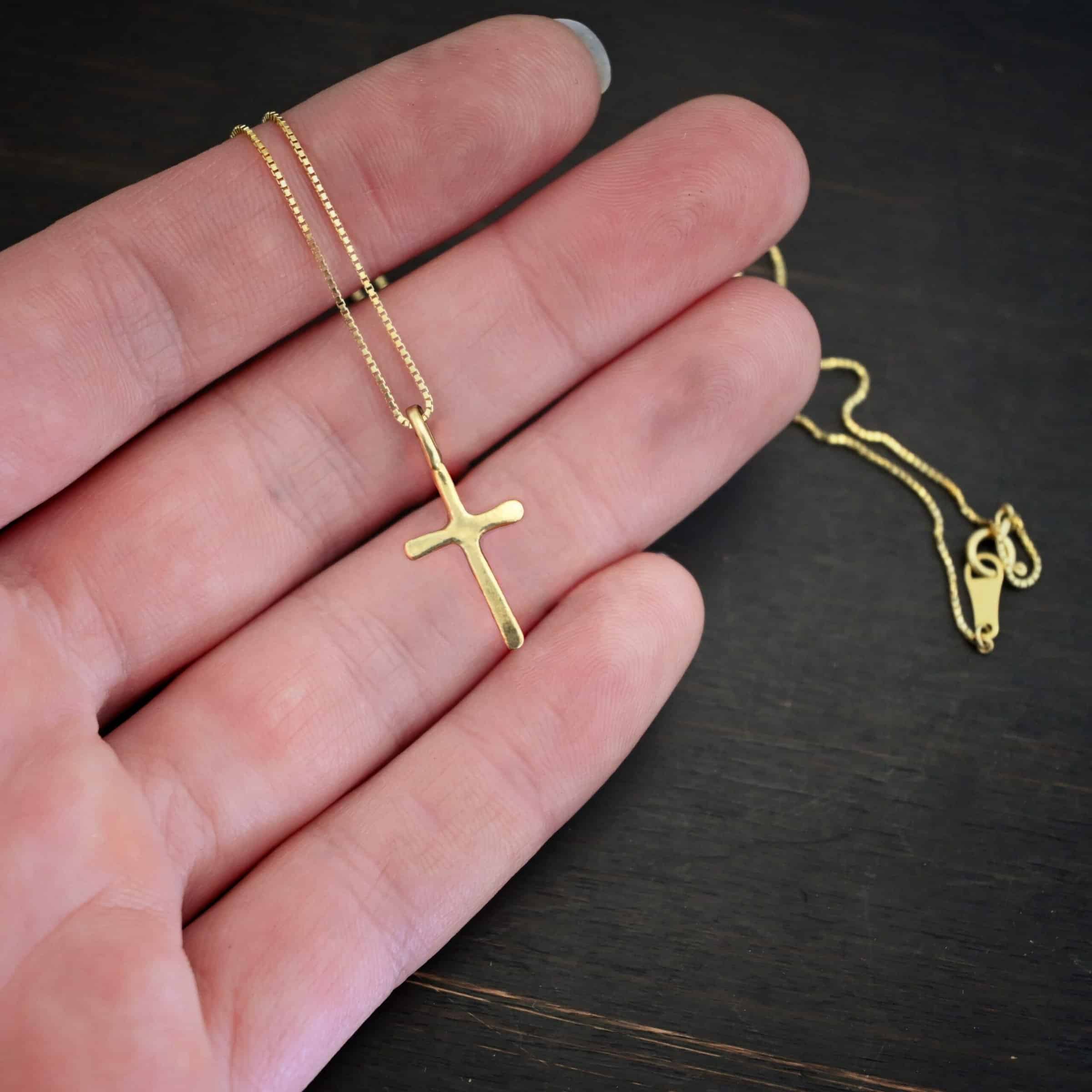 Divine Necklace Photo of the "Divine" Necklace. Made out of 18k gold. There is a small sized cross that is flat and smooth. It has a loop soldered to the top of it. There is a chain that goes through the loop of the cross that is also gold. Necklace is lying against the fingertips of a hand to demonstrate size.