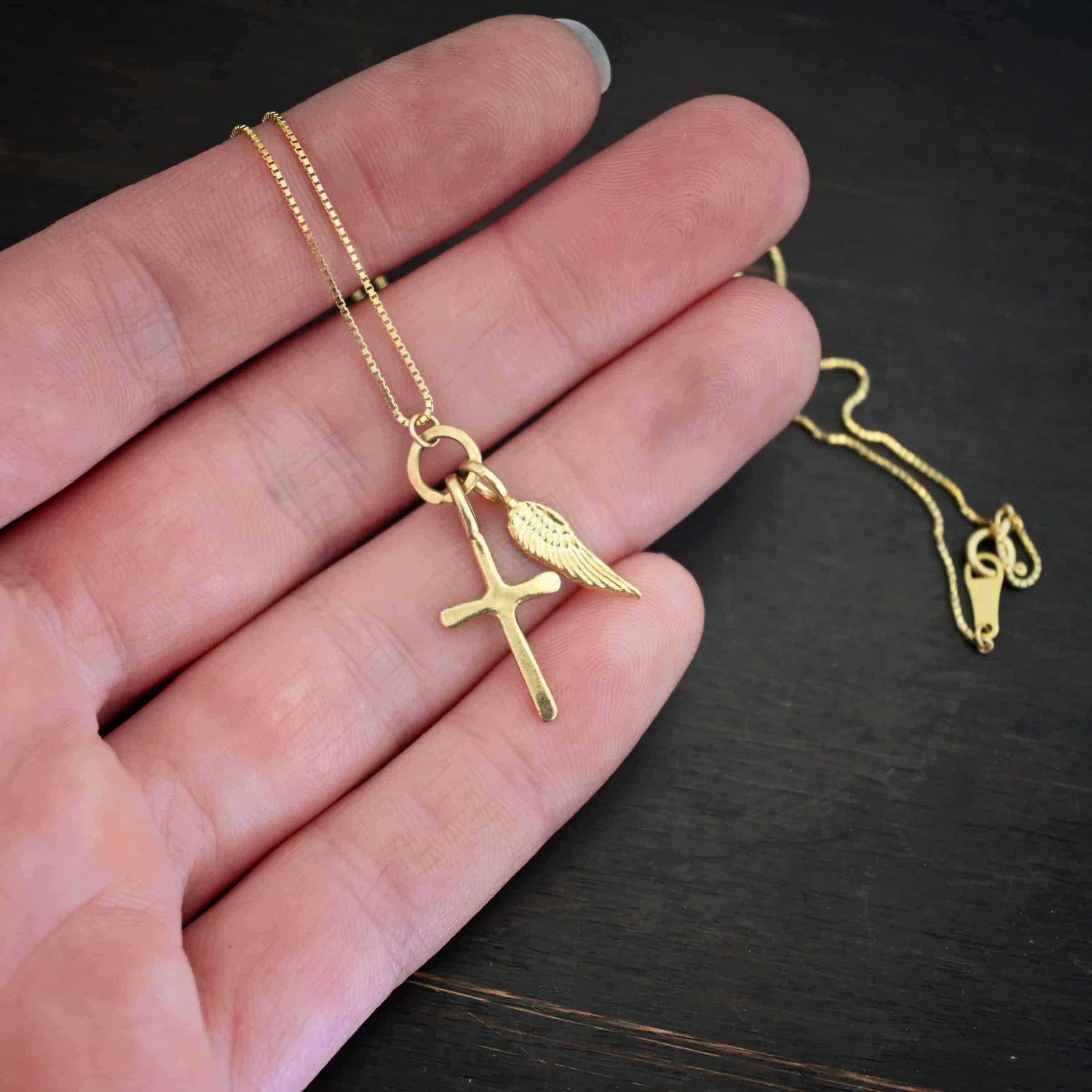 Divine Necklace Photo of the "Divine" Necklace. Made out of 18k gold. There is a small sized cross that is flat and smooth. It has a loop soldered to the top of it. There is a small sized angel wing pendant next to it, also with a soldered loop on the top. There is a chain that goes through the top of the necklace that is also gold. Necklace is lying against the fingertips of a hand to demonstrate size.