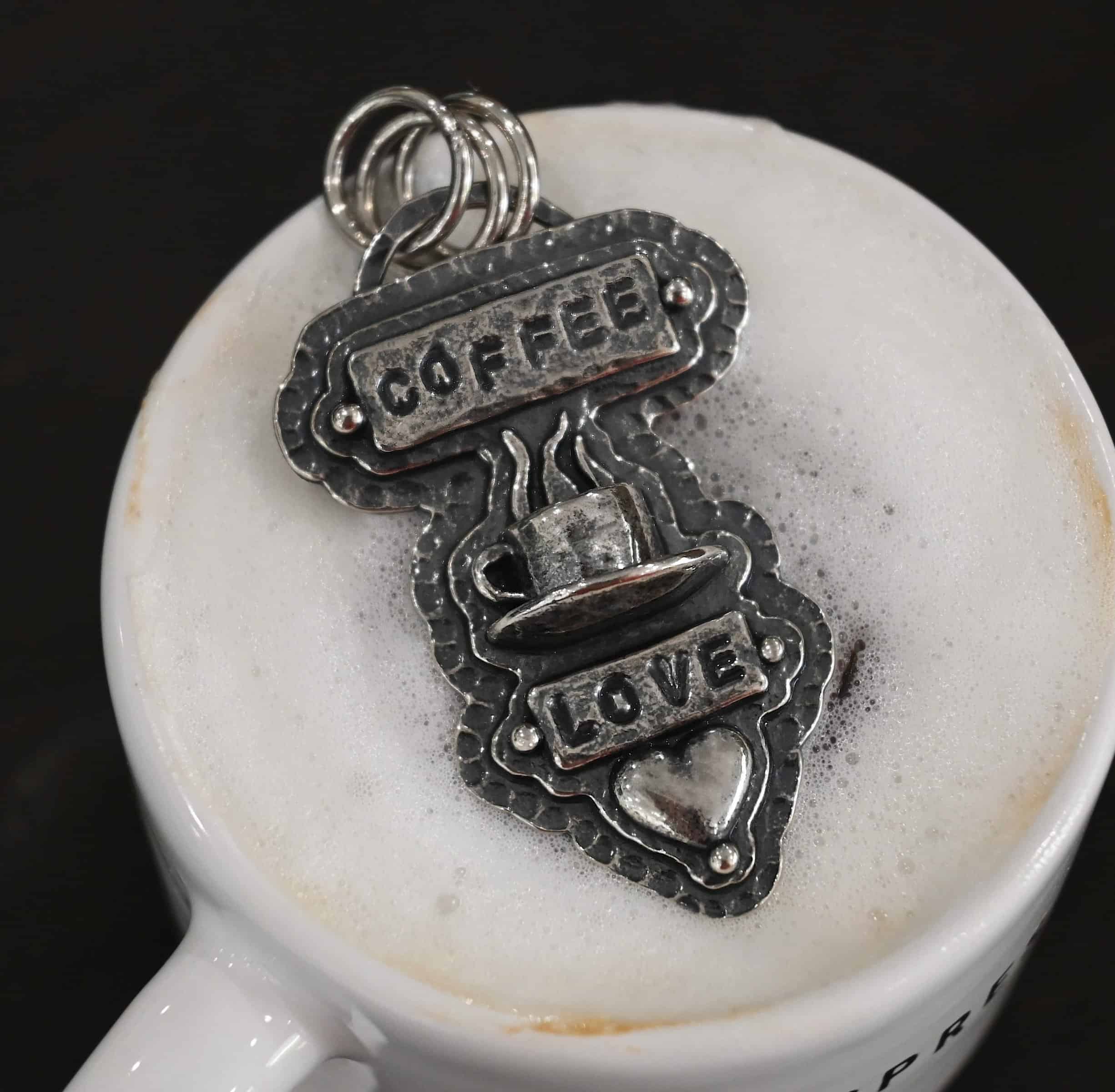 True Addiction Coffee Necklace Photo of the "True Addiction" Coffee Necklace. The pendant has two layers of sterling silver, both of which are hammered. The top layer is smaller than the bottom layer, so that both are noticeable. The top layer has a rectangle bar of silver soldered to it that has the word "Coffee" stamped into it. On each end of the bar is a sterling silver ball. Beneath the bar, soldered to the top layer is a coffee cup charm sitting on a saucer with steam waves coming out of the top of it. Beneath the saucer of the cup is another smaller rectangle bar on the top layer that has "love" stamped into it. There is a silver ball on each end of the rectangle. Beneath the rectangle is a heart charm that is soldered. Beneath the heart is a silver ball on the tip of the heart. The top of the necklace, on the back, has two loops, one loop on each side. Attached to each loop is the chain of the necklace. Necklace is lying on the top of coffee with foam on it.