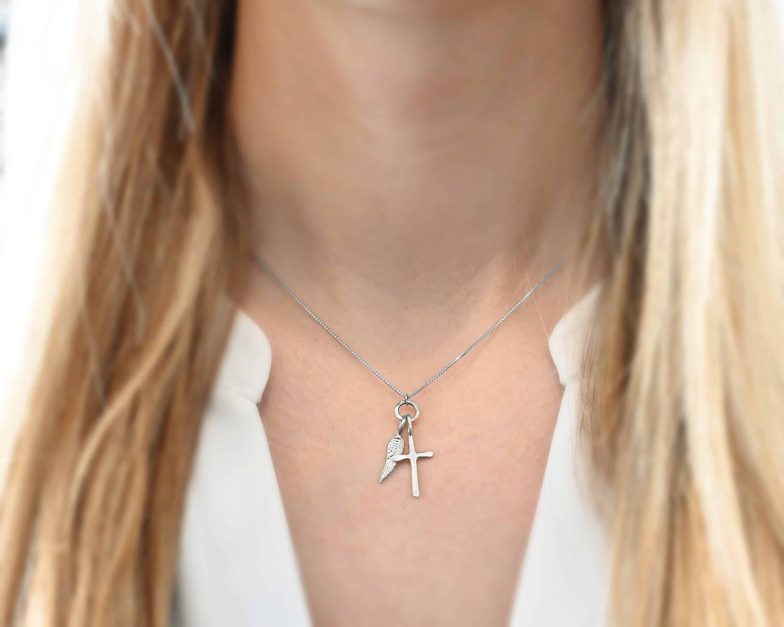 Divine Necklacev Photo of the "Divine" Necklace. Made out of sterling silver. There is a small sized cross that is flat and smooth. It has a loop soldered to the top of it. There is a small sized angel wing pendant next to it, also with a soldered loop on the top. There is a chain that goes through the top of the necklace that is also silver. Necklace is worn by a model to demonstrate size.