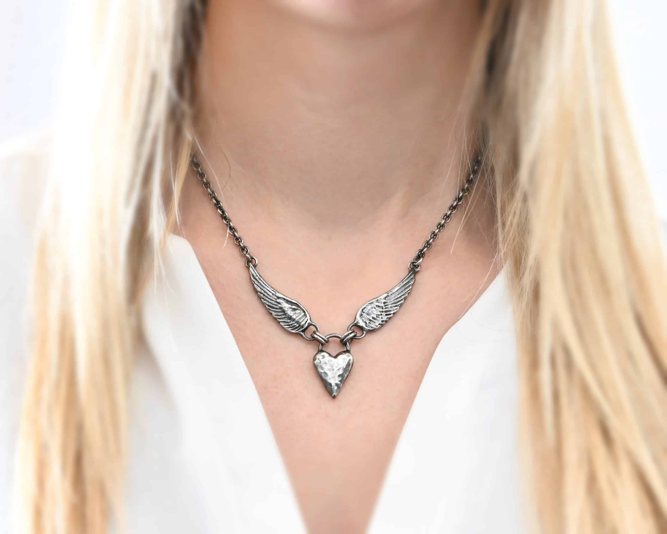 Courage Necklace Photo of the "Courage" Necklace. Made out of sterling silver. There is a thick, solid sterling silver heart that is a medium size for the pendant. It is hammered and oxidized. There is a thick semicircle that is soldered on both sides of the heart on the top. There are four jump rings attached to the semicircle, two on each side. Attached to the jump rings on each side are two angel wings that face opposite directions. The tip of each wing has a loop soldered to the back of them. Attached to the loop on the back of each wing is the chain of the necklace. Necklace is worn by a model and shown up close to demonstrate size.