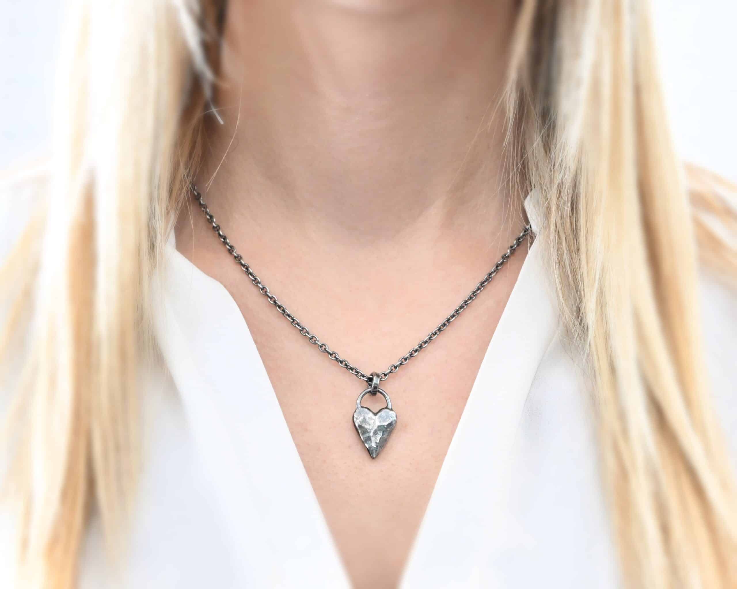 Photo of the "Heart Throb" Necklace. Made out of sterling silver. There is a thick, solid sterling silver heart that is a medium size for the pendant. It is hammered and oxidized. There is a thick semicircle that is soldered on both sides of the heart on the top. Attached to the semicircle is a thick jump ring that the chain of the necklace goes through. Necklace is worn by a model who is wearing a white v-neck shirt.