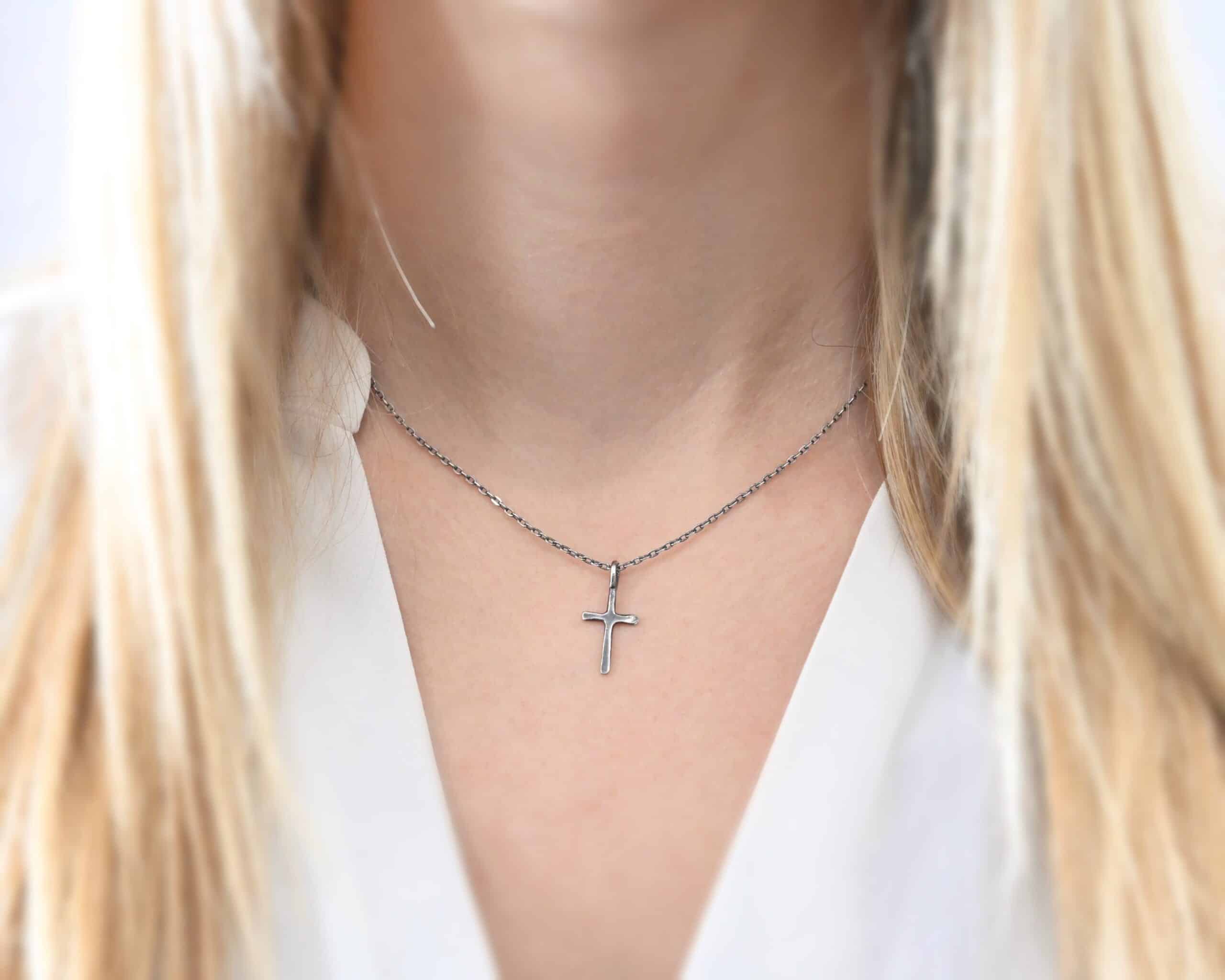 Redeemer Necklace Photo of the "Redeemer" Necklace. Made out of sterling silver and oxidized. Necklace has a very small cross that is hammered flat, but smooth, with a loop soldered to the top of it. The chain of the necklace is oxidized. Necklace is worn by a model who is wearing a white v-neck shirt.