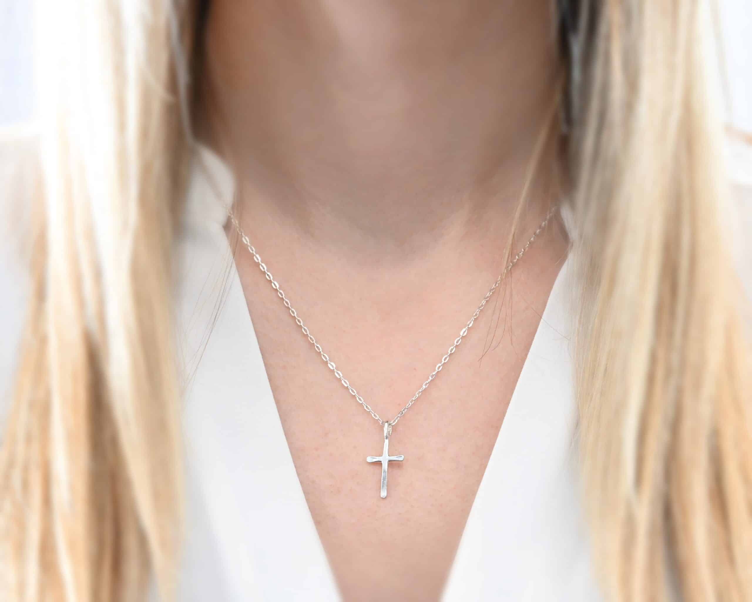Redeemer Necklace Photo of the "Redeemer" Necklace. Made out of sterling silver and shiny. Necklace has a very small cross that is hammered flat, but smooth, with a loop soldered to the top of it. The chain of the necklace is shiny. Necklace is worn by a model who is wearing a white v-neck shirt.