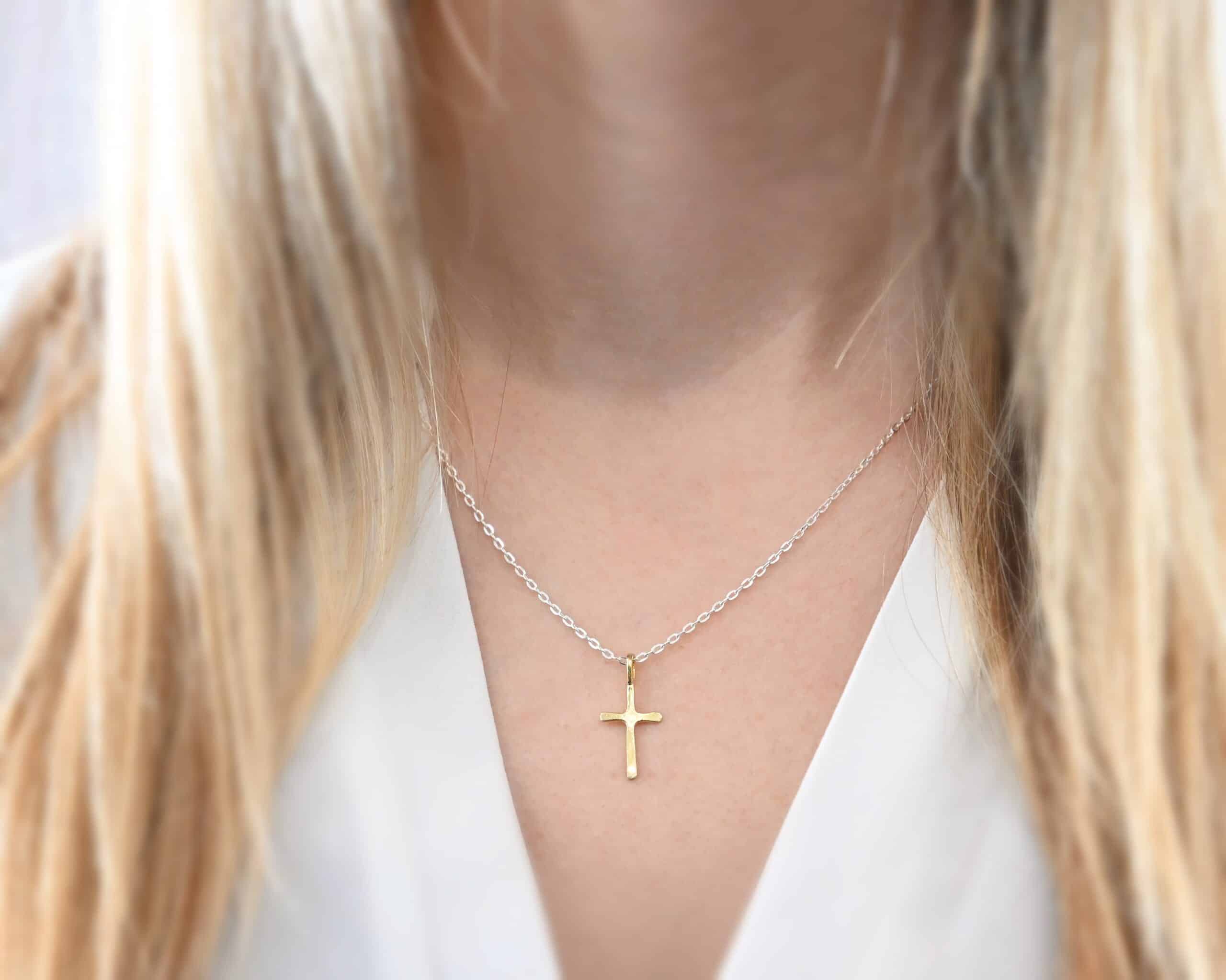 Redeemer Necklace Photo of the "Redeemer" Necklace. Made out of 18k gold. Necklace has a very small cross that is hammered flat, but smooth, with a loop soldered to the top of it. The chain of the necklace is shiny. Necklace is worn by a model who is wearing a white v-neck shirt.