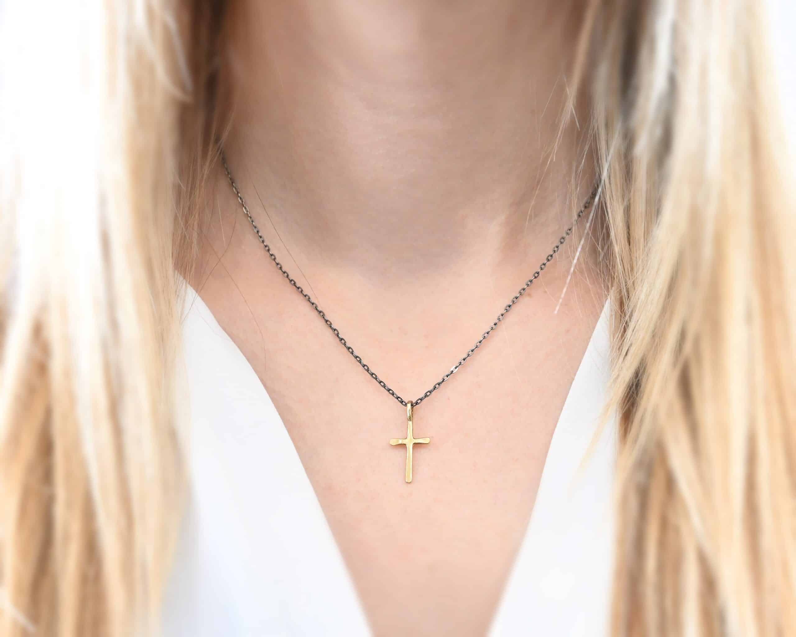 Redeemer Necklace Photo of the "Redeemer" Necklace. Made out of 18k gold. Necklace has a very small cross that is hammered flat, but smooth, with a loop soldered to the top of it. The chain of the necklace is oxidized. Necklace is worn by a model who is wearing a white v-neck shirt.