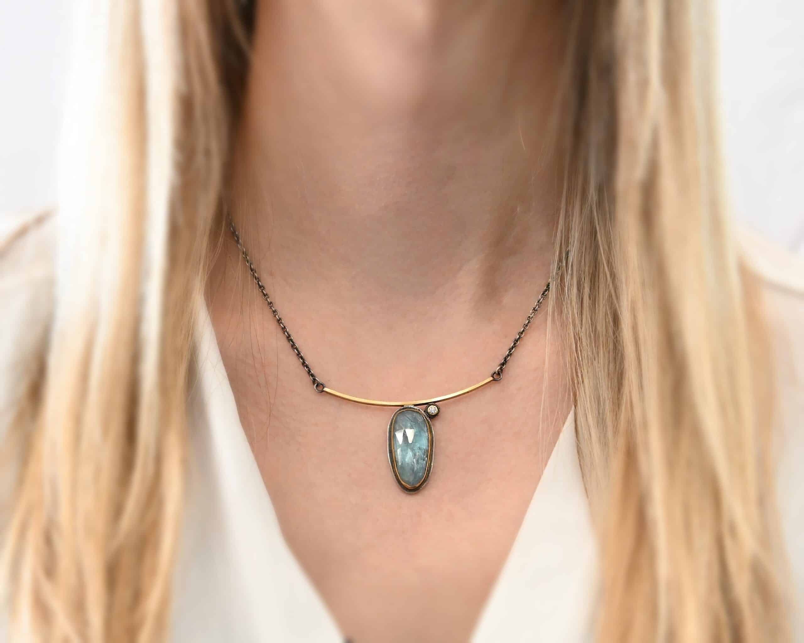 Photo of the "Day Dream" Necklace. Made out of sterling silver, gold, kyanite, and moissanite. There is an oval shaped kyanite set vertically in gold that is soldered to the bottom of a curved gold bar that is smooth. The kyanite has a silver back behind it to keep the stone in place. There is a small moissanite on the top right side of the kyanite. The moissanite is also soldered to the bar of the necklace. It is set in gold with silver behind it. The bar of gold has a silver loop on each end that each end of the chain attaches to. Necklace is worn by a model to demonstrate size.