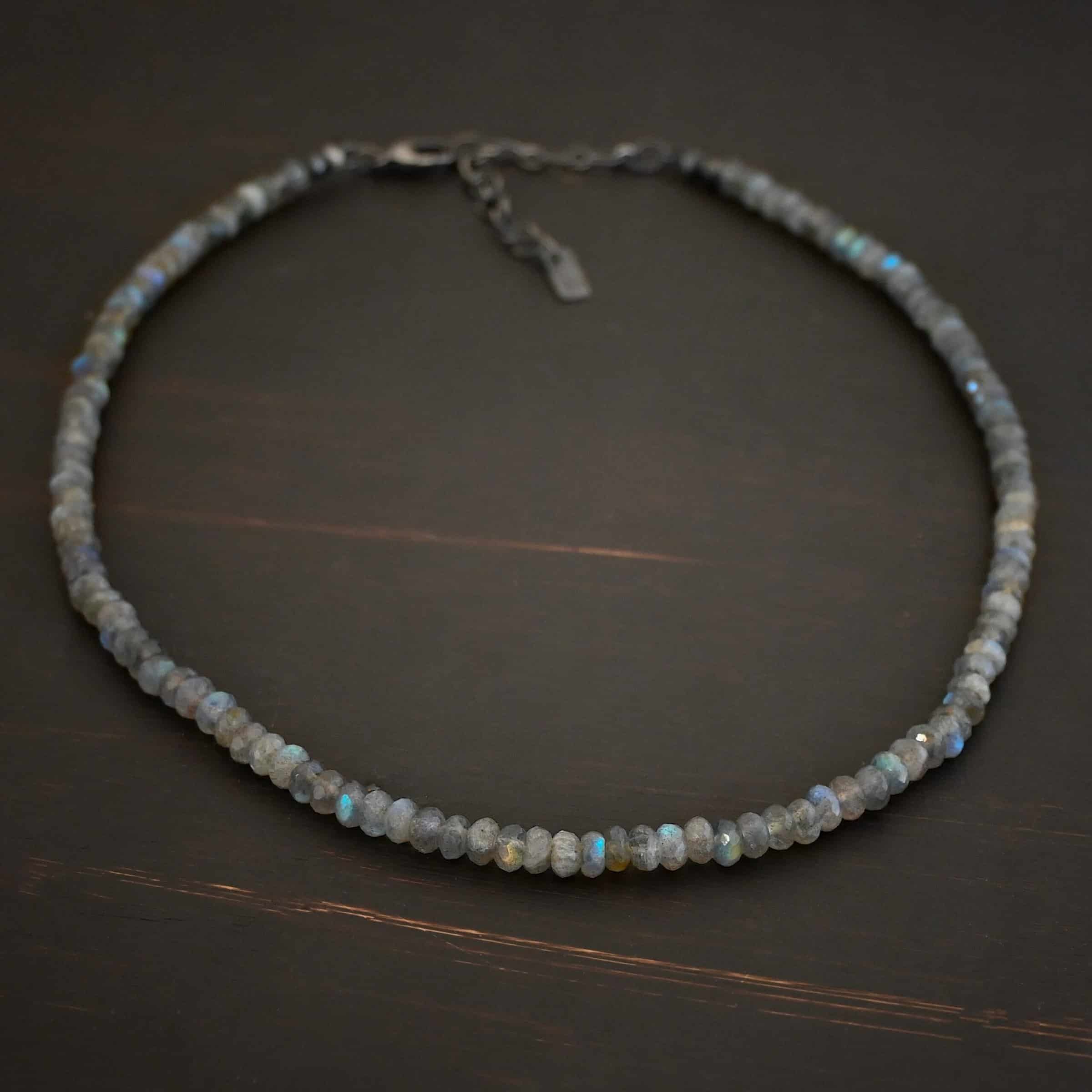 Lucy Labradorite Necklace Photo of the "Lucy Labradorite" Necklace. It is made of labradorite beads strung tightly together on silver wire. There are silver beads and crimps on the end of the necklace, and a silver clasp. Necklace is lying against a dark background.