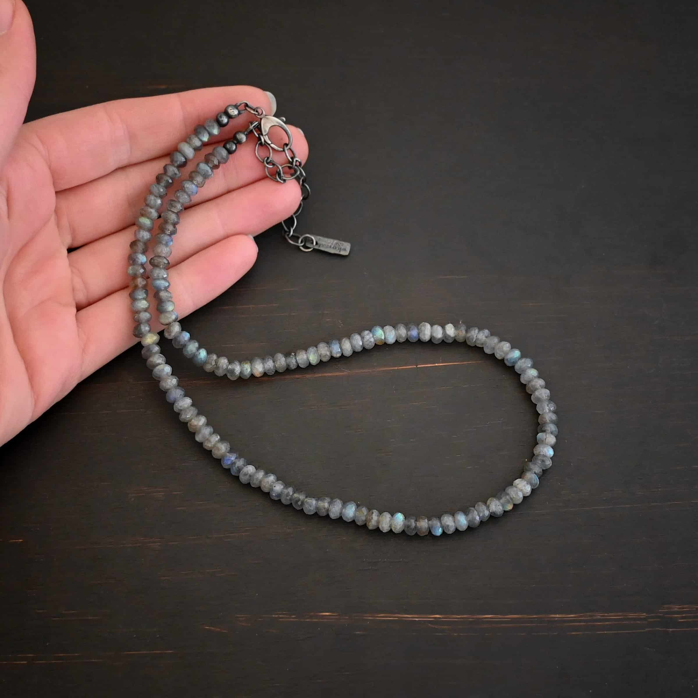 Lucy Labradorite Necklace Photo of the "Lucy Labradorite" Necklace. It is made of labradorite beads strung tightly together on silver wire. There are silver beads and crimps on the end of the necklace, and a silver clasp. Necklace is lying against the fingertips of a hand.