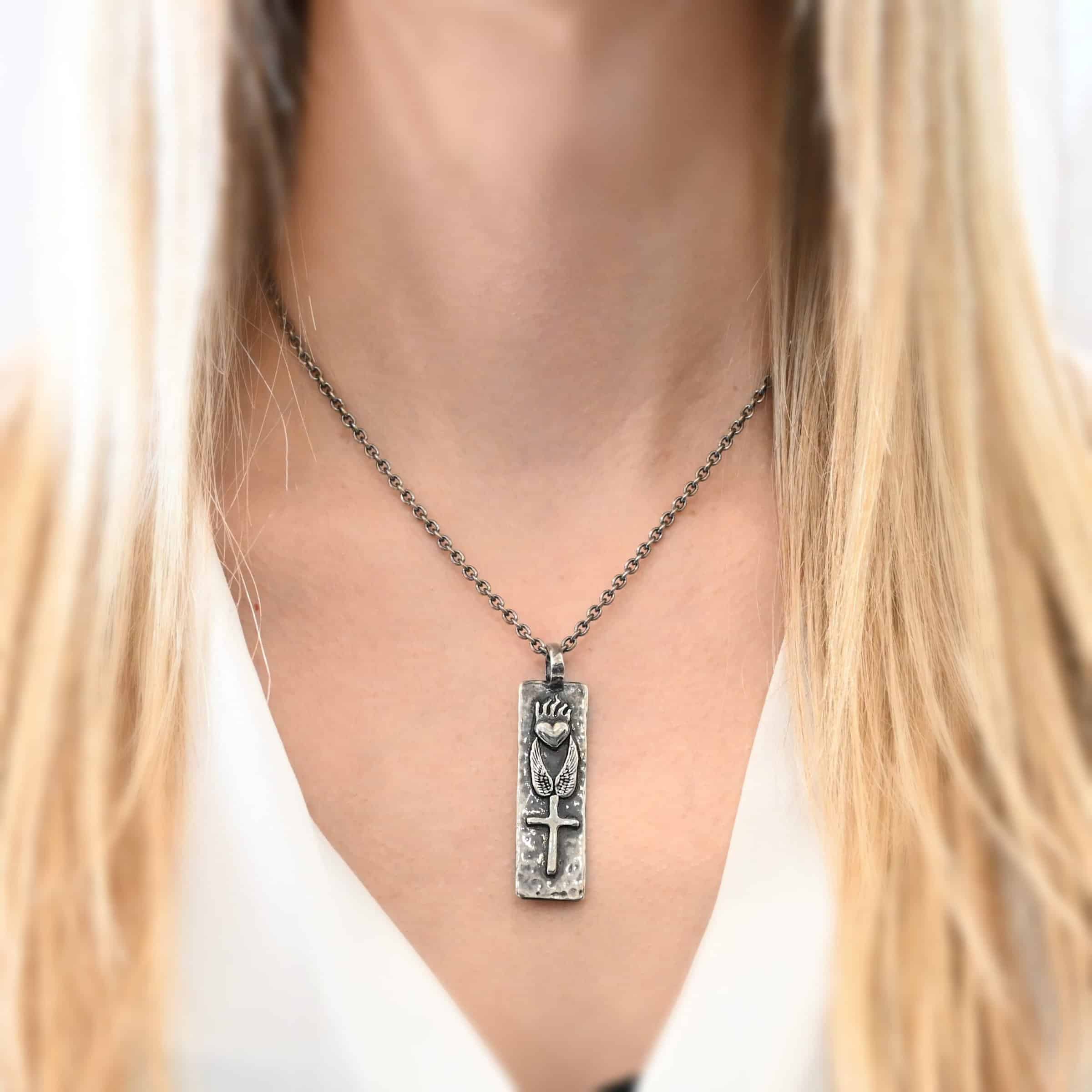 Protect Necklace Photo of the "Protect" Necklace. Necklace is made out of silver. The pendant is a rectangle shaped silver tag that hangs vertically. It is large. On the top of the rectangle is a silver heart with flames dancing across the top of it. Beneath this are two silver angel wings, one on each side of the heart. Beneath the angel wings in the center of them and on the bottom of the rectangle pendant is a silver cross soldered to the pendant. There is a thick jump ring soldered to the top of the pendant that the chain of the necklace goes through. Necklace is worn by a model to demonstrate size.
