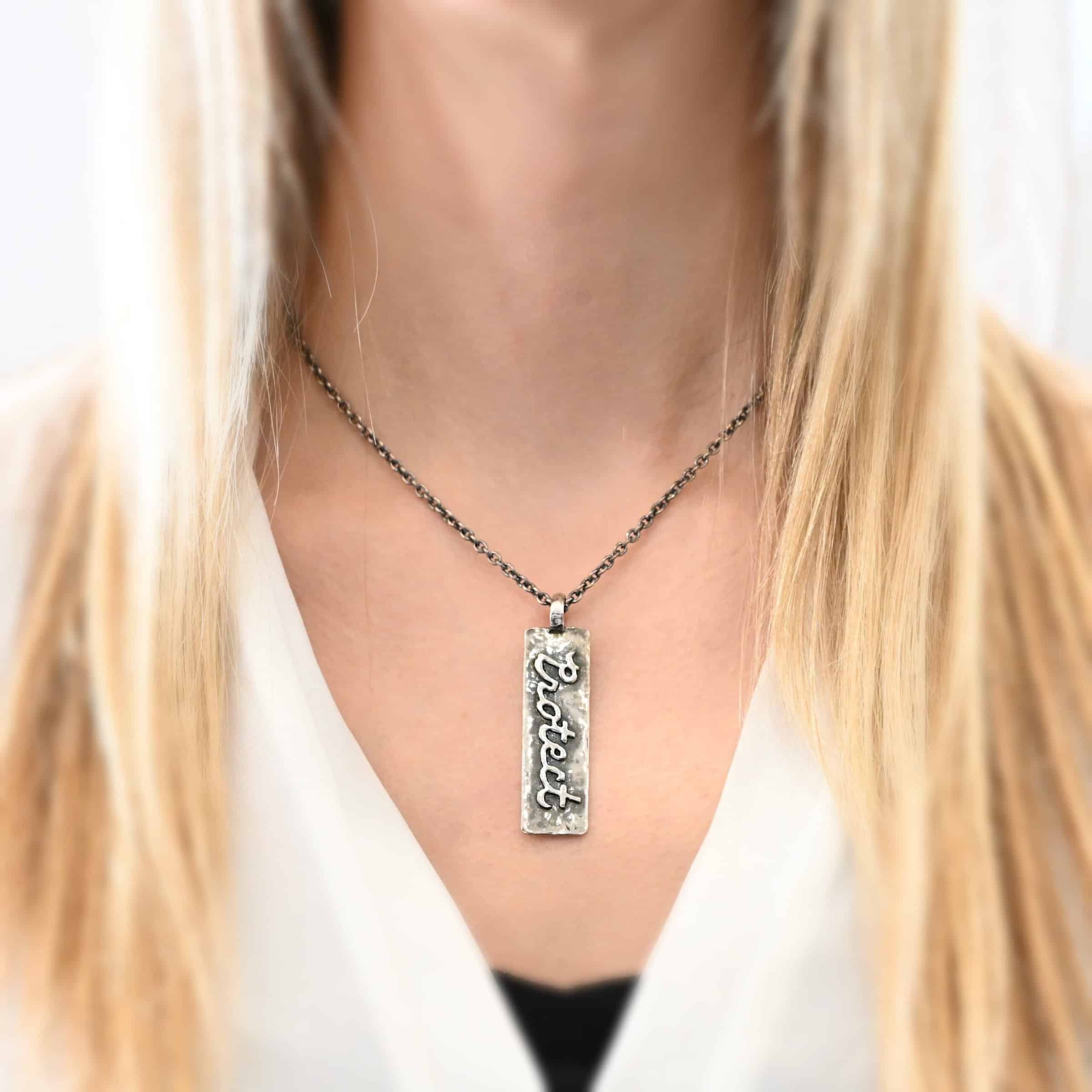 Protect Necklace Photo of the "Protect" Necklace. Necklace is made out of silver. The pendant is a rectangle shaped silver tag that hangs vertically. It is large. It has the word "Protect" soldered across it in cursive. There is a thick jump ring soldered to the top of the pendant that the chain of the necklace goes through. Necklace is worn by a model to demonstrate size.
