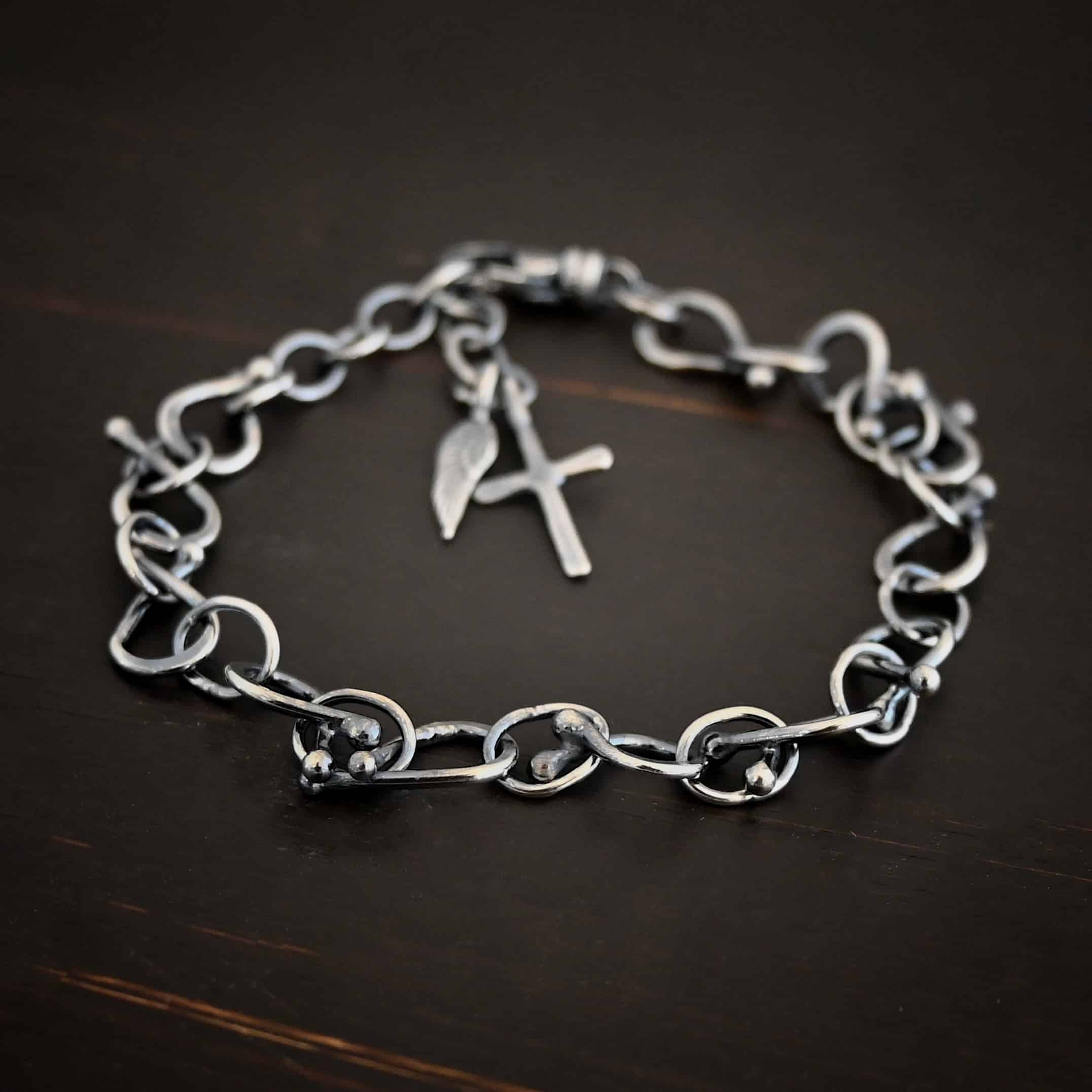 Loretta Bracelet Photo of the "Loretta" Bracelet. Made out of sterling silver. The bracelet is made out of pear shaped. The top of each link has two silver balls, one for each end at the top. Between each teardrop link is a circle link of silver. There is a cross and angel wing charm on the bracelet, and bracelet has a lobster claw clasp. Bracelet is lying against a dark background.