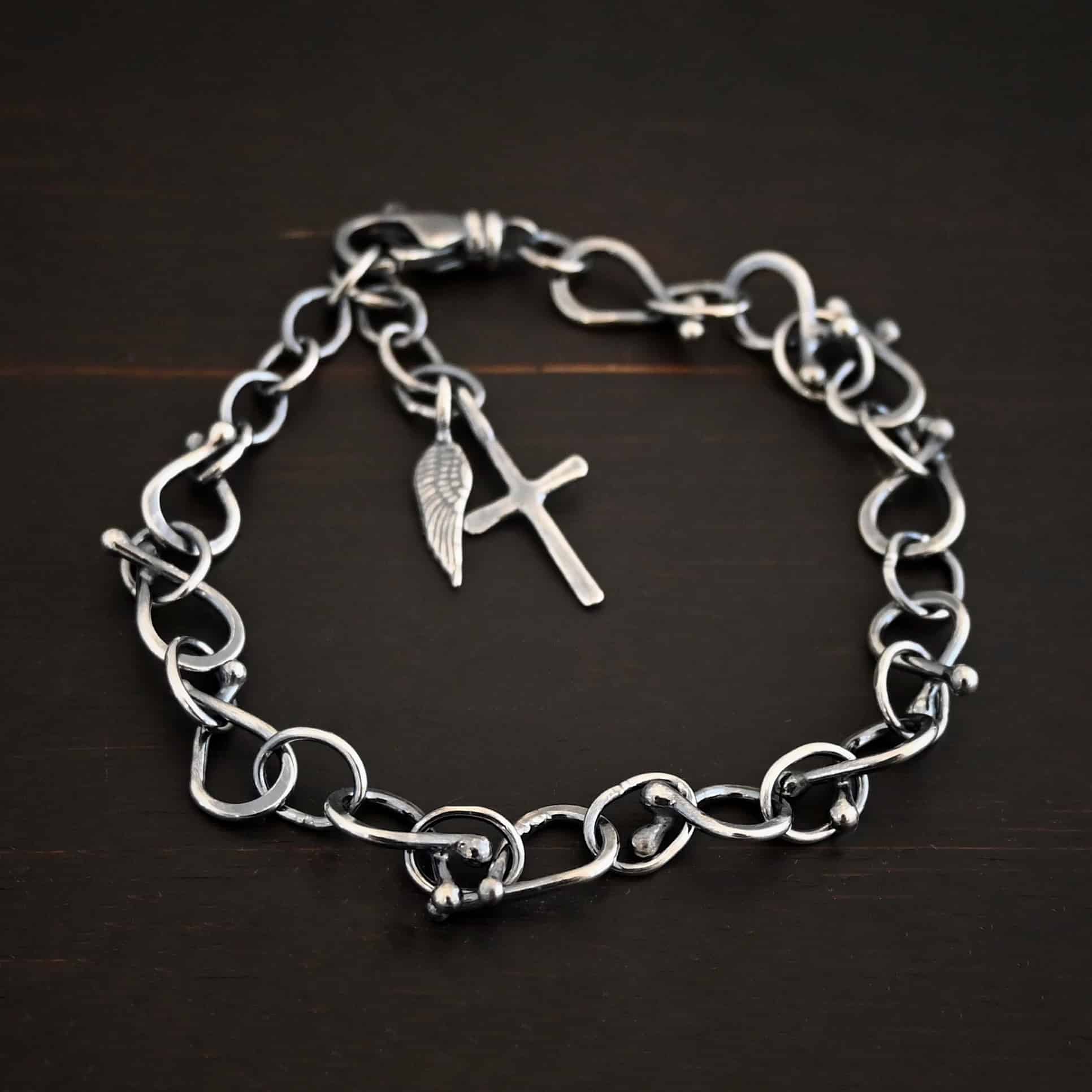 Loretta Bracelet Photo of the "Loretta" Bracelet. Made out of sterling silver. The bracelet is made out of pear shaped. The top of each link has two silver balls, one for each end at the top. Between each teardrop link is a circle link of silver. There is a cross and angel wing charm on the bracelet, and bracelet has a lobster claw clasp. Bracelet is lying against a dark background.