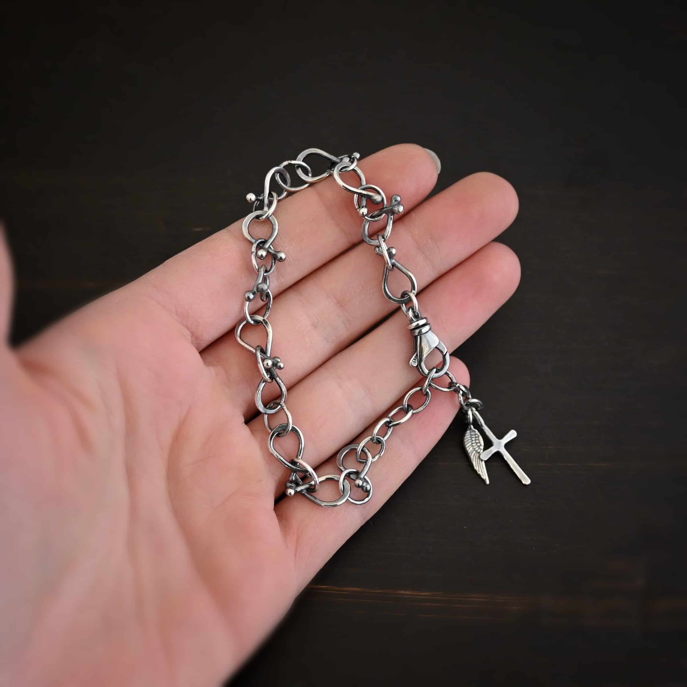 Loretta Bracelet Photo of the "Loretta" Bracelet. Made out of sterling silver. The bracelet is made out of pear shaped. The top of each link has two silver balls, one for each end at the top. Between each teardrop link is a circle link of silver. There is a cross and angel wing charm on the bracelet, and bracelet has a lobster claw clasp. Bracelet is lying against the fingertips of a hand to demonstrate size.