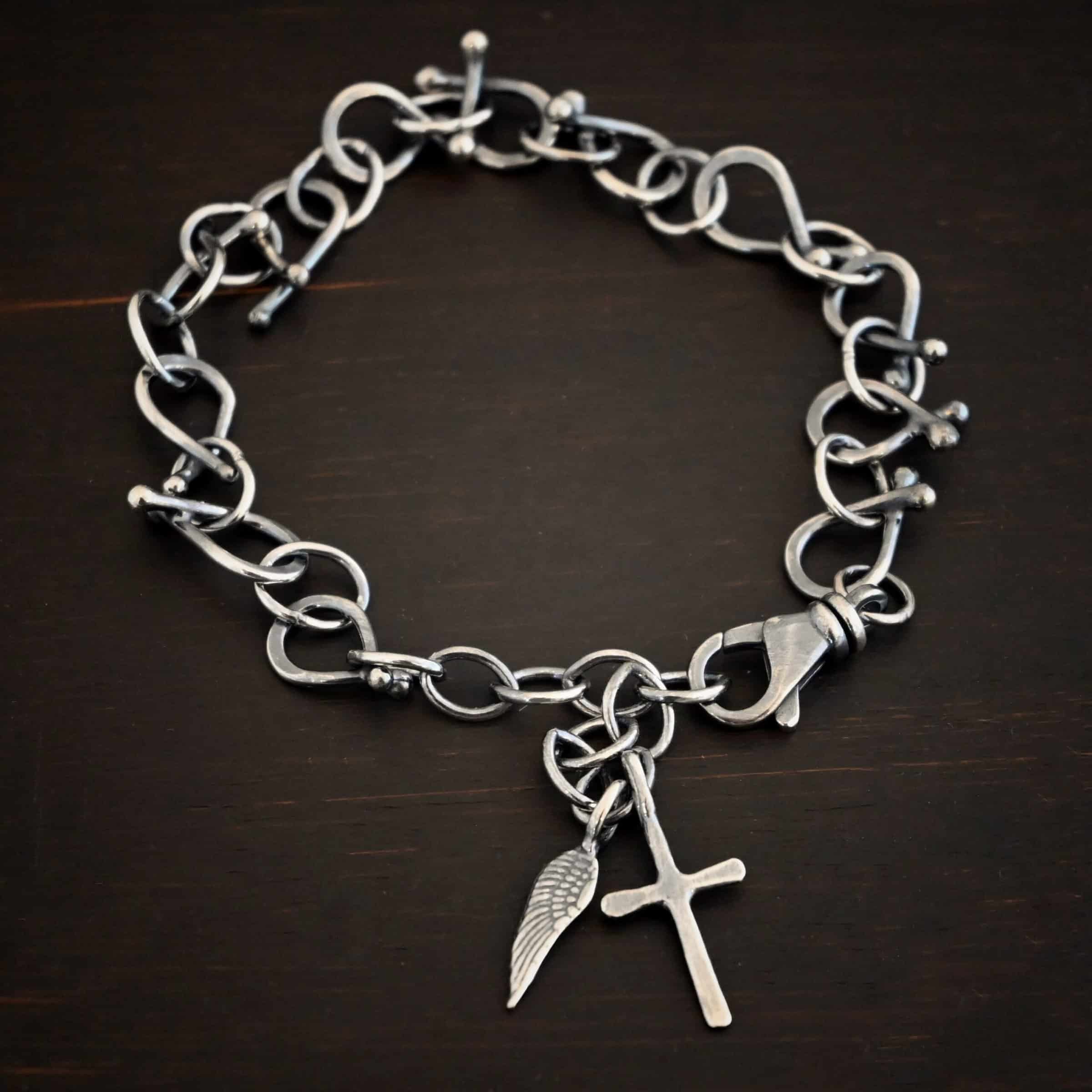 Loretta Bracelet Photo of the "Loretta" Bracelet. Made out of sterling silver. The bracelet is made out of pear shaped. The top of each link has two silver balls, one for each end at the top. Between each teardrop link is a circle link of silver. There is a cross and angel wing charm on the bracelet, and bracelet has a lobster claw clasp. Bracelet is lying against a dark background.