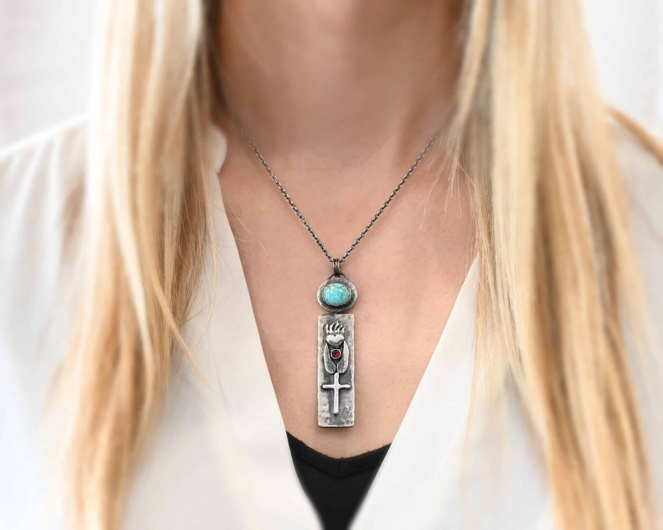 Photo of the "Sacred Heart" Necklace. Necklace is made out of silver, garnet, and turquoise. The pendant is a rectangle shaped silver tag that hangs vertically. It is large. On the top of the rectangle is a silver heart with flames dancing across the top of it. Beneath the heart on the bottom tip of it is a circular small garnet. Beneath this are two silver angel wings, one on each side of the garnet surrounding it. Beneath the angel wings in the center of them and on the bottom of the rectangle pendant is a silver cross soldered to the pendant. There is an oval shaped turquoise soldered to the top of the rectangular pendant. The silver around the turquoise is hammered and oxidized. There is a loop on the back of the turquoise with three jump rings going through it. The chain of the necklace goes through the jump rings. Necklace is worn by a model to demonstrate size.