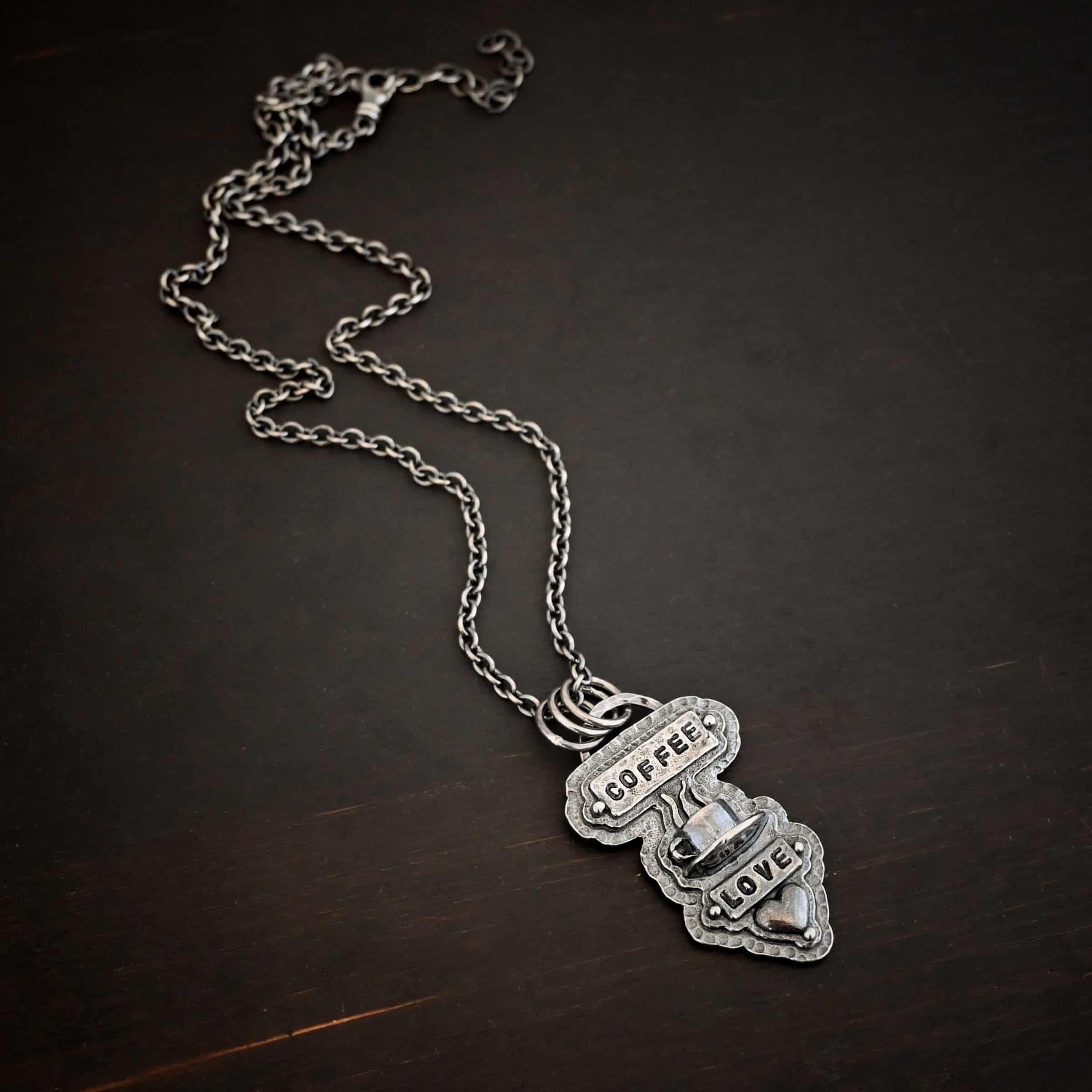 True Addiction Coffee Necklace Photo of the "True Addiction" Coffee Necklace. The pendant has two layers of sterling silver, both of which are hammered. The top layer is smaller than the bottom layer, so that both are noticeable. The top layer has a rectangle bar of silver soldered to it that has the word "Coffee" stamped into it. On each end of the bar is a sterling silver ball. Beneath the bar, soldered to the top layer is a coffee cup charm sitting on a saucer with steam waves coming out of the top of it. Beneath the saucer of the cup is another smaller rectangle bar on the top layer that has "love" stamped into it. There is a silver ball on each end of the rectangle. Beneath the rectangle is a heart charm that is soldered. Beneath the heart is a silver ball on the tip of the heart. The top of the necklace, on the back, has two loops, one loop on each side. Attached to each loop is the chain of the necklace. Necklace is lying against a dark background.