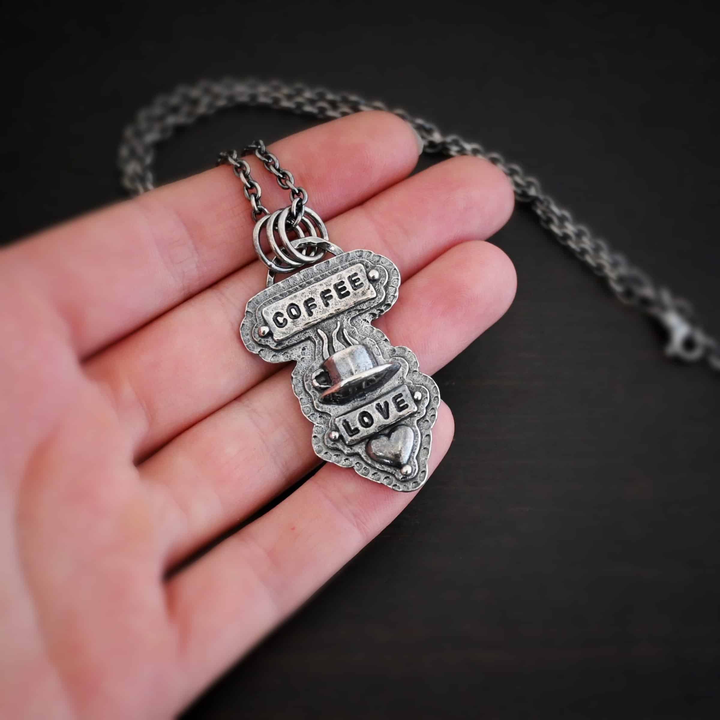 True Addiction Coffee Necklace Photo of the "True Addiction" Coffee Necklace. The pendant has two layers of sterling silver, both of which are hammered. The top layer is smaller than the bottom layer, so that both are noticeable. The top layer has a rectangle bar of silver soldered to it that has the word "Coffee" stamped into it. On each end of the bar is a sterling silver ball. Beneath the bar, soldered to the top layer is a coffee cup charm sitting on a saucer with steam waves coming out of the top of it. Beneath the saucer of the cup is another smaller rectangle bar on the top layer that has "love" stamped into it. There is a silver ball on each end of the rectangle. Beneath the rectangle is a heart charm that is soldered. Beneath the heart is a silver ball on the tip of the heart. The top of the necklace, on the back, has two loops, one loop on each side. Attached to each loop is the chain of the necklace. Necklace is lying against the fingertips of a hand to demonstrate size.