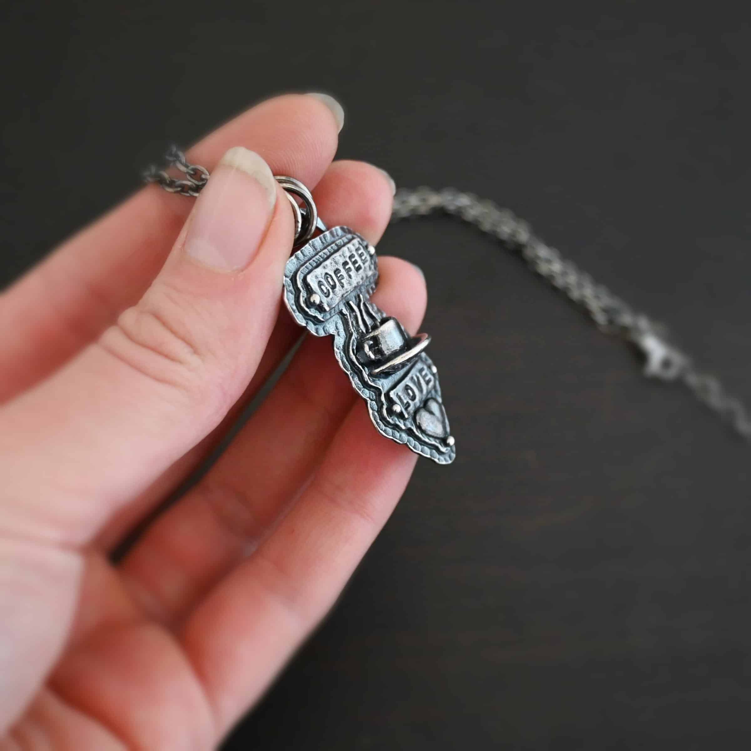 True Addiction Coffee Necklace Photo of the "True Addiction" Coffee Necklace. The pendant has two layers of sterling silver, both of which are hammered. The top layer is smaller than the bottom layer, so that both are noticeable. The top layer has a rectangle bar of silver soldered to it that has the word "Coffee" stamped into it. On each end of the bar is a sterling silver ball. Beneath the bar, soldered to the top layer is a coffee cup charm sitting on a saucer with steam waves coming out of the top of it. Beneath the saucer of the cup is another smaller rectangle bar on the top layer that has "love" stamped into it. There is a silver ball on each end of the rectangle. Beneath the rectangle is a heart charm that is soldered. Beneath the heart is a silver ball on the tip of the heart. The top of the necklace, on the back, has two loops, one loop on each side. Attached to each loop is the chain of the necklace. Necklace is lying against the fingertips of a hand to demonstrate size.