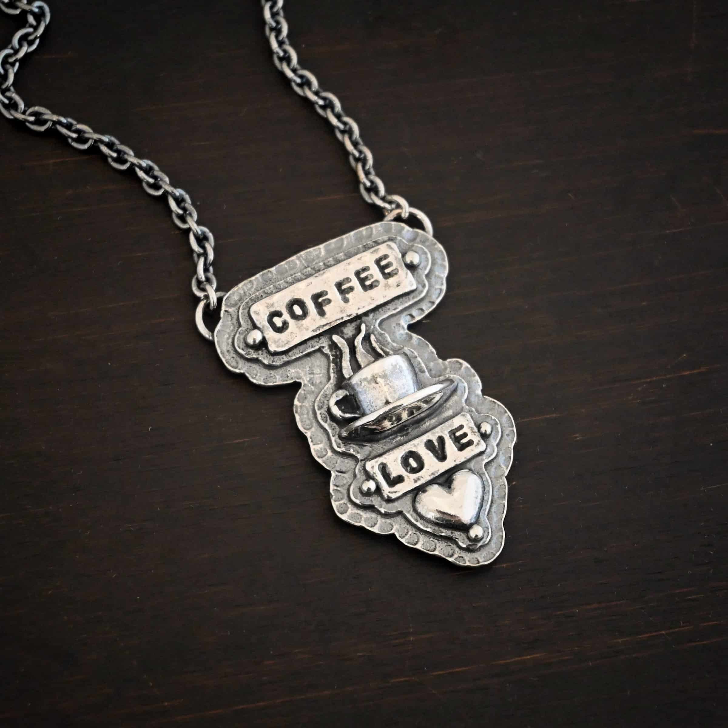 True Addiction Coffee Necklace Photo of the "True Addiction" Coffee Necklace. The pendant has two layers of sterling silver, both of which are hammered. The top layer is smaller than the bottom layer, so that both are noticeable. The top layer has a rectangle bar of silver soldered to it that has the word "Coffee" stamped into it. On each end of the bar is a sterling silver ball. Beneath the bar, soldered to the top layer is a coffee cup charm sitting on a saucer with steam waves coming out of the top of it. Beneath the saucer of the cup is another smaller rectangle bar on the top layer that has "love" stamped into it. There is a silver ball on each end of the rectangle. Beneath the rectangle is a heart charm that is soldered. Beneath the heart is a silver ball on the tip of the heart. The top of the necklace, on the back, has two loops, one loop on each side. Attached to each loop is the chain of the necklace. Necklace is lying against a dark background.
