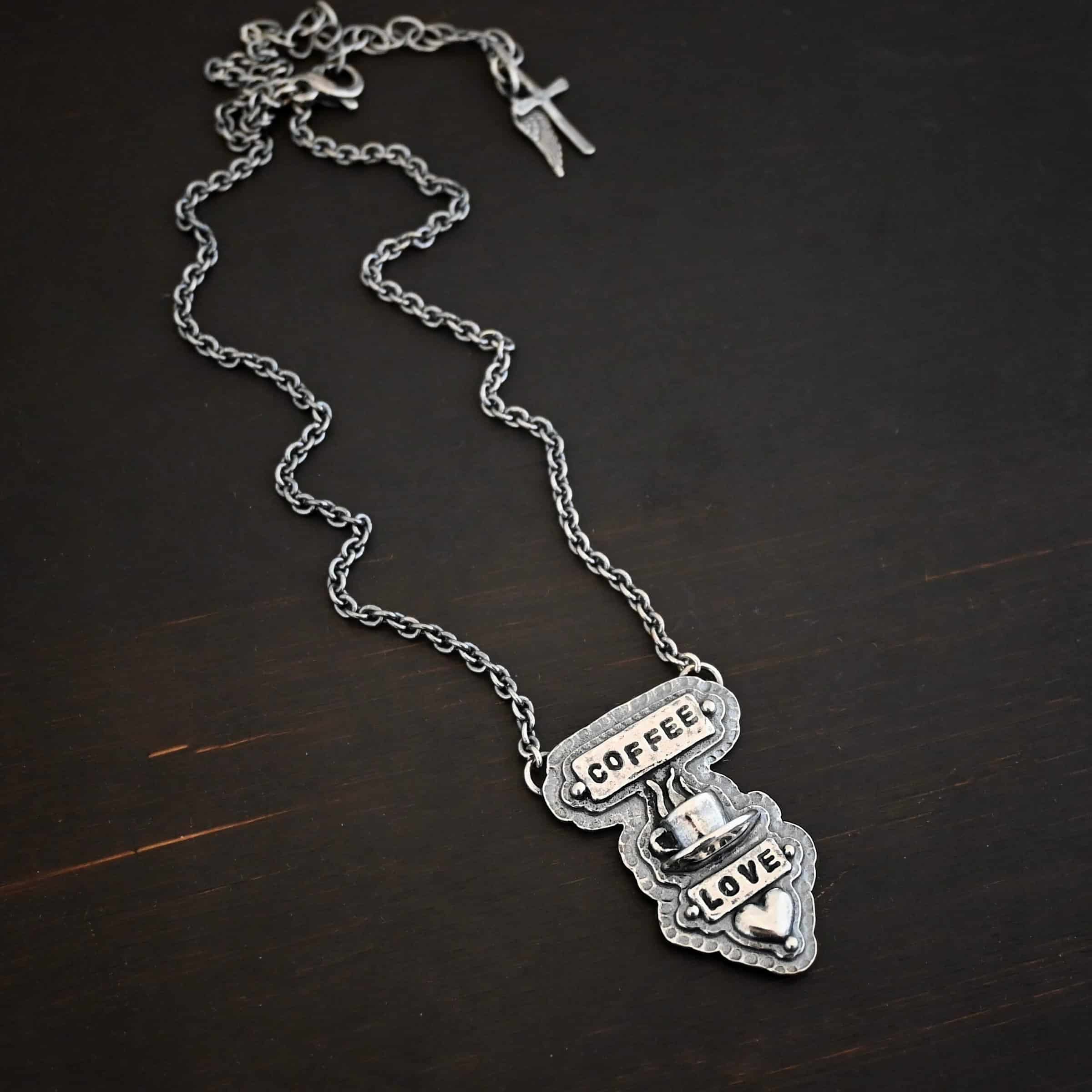 True Addiction Coffee Necklace Photo of the "True Addiction" Coffee Necklace. The pendant has two layers of sterling silver, both of which are hammered. The top layer is smaller than the bottom layer, so that both are noticeable. The top layer has a rectangle bar of silver soldered to it that has the word "Coffee" stamped into it. On each end of the bar is a sterling silver ball. Beneath the bar, soldered to the top layer is a coffee cup charm sitting on a saucer with steam waves coming out of the top of it. Beneath the saucer of the cup is another smaller rectangle bar on the top layer that has "love" stamped into it. There is a silver ball on each end of the rectangle. Beneath the rectangle is a heart charm that is soldered. Beneath the heart is a silver ball on the tip of the heart. The top of the necklace, on the back, has two loops, one loop on each side. Attached to each loop is the chain of the necklace. Necklace is lying against a dark background.