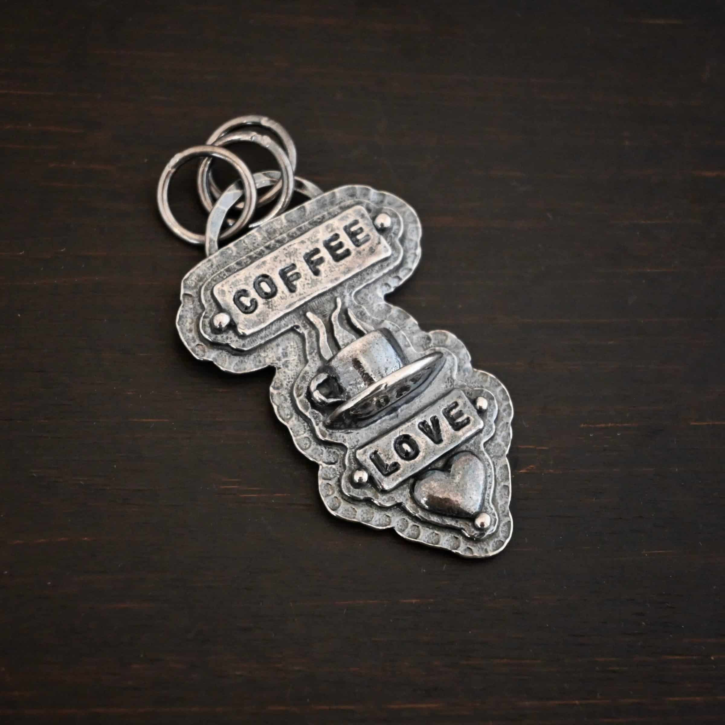 True Addiction Coffee Necklace Photo of the "True Addiction" Coffee Necklace. The pendant has two layers of sterling silver, both of which are hammered. The top layer is smaller than the bottom layer, so that both are noticeable. The top layer has a rectangle bar of silver soldered to it that has the word "Coffee" stamped into it. On each end of the bar is a sterling silver ball. Beneath the bar, soldered to the top layer is a coffee cup charm sitting on a saucer with steam waves coming out of the top of it. Beneath the saucer of the cup is another smaller rectangle bar on the top layer that has "love" stamped into it. There is a silver ball on each end of the rectangle. Beneath the rectangle is a heart charm that is soldered. Beneath the heart is a silver ball on the tip of the heart. The top of the necklace, on the back, has two loops, one loop on each side. Attached to each loop is the chain of the necklace. Necklace is lying against a dark background.