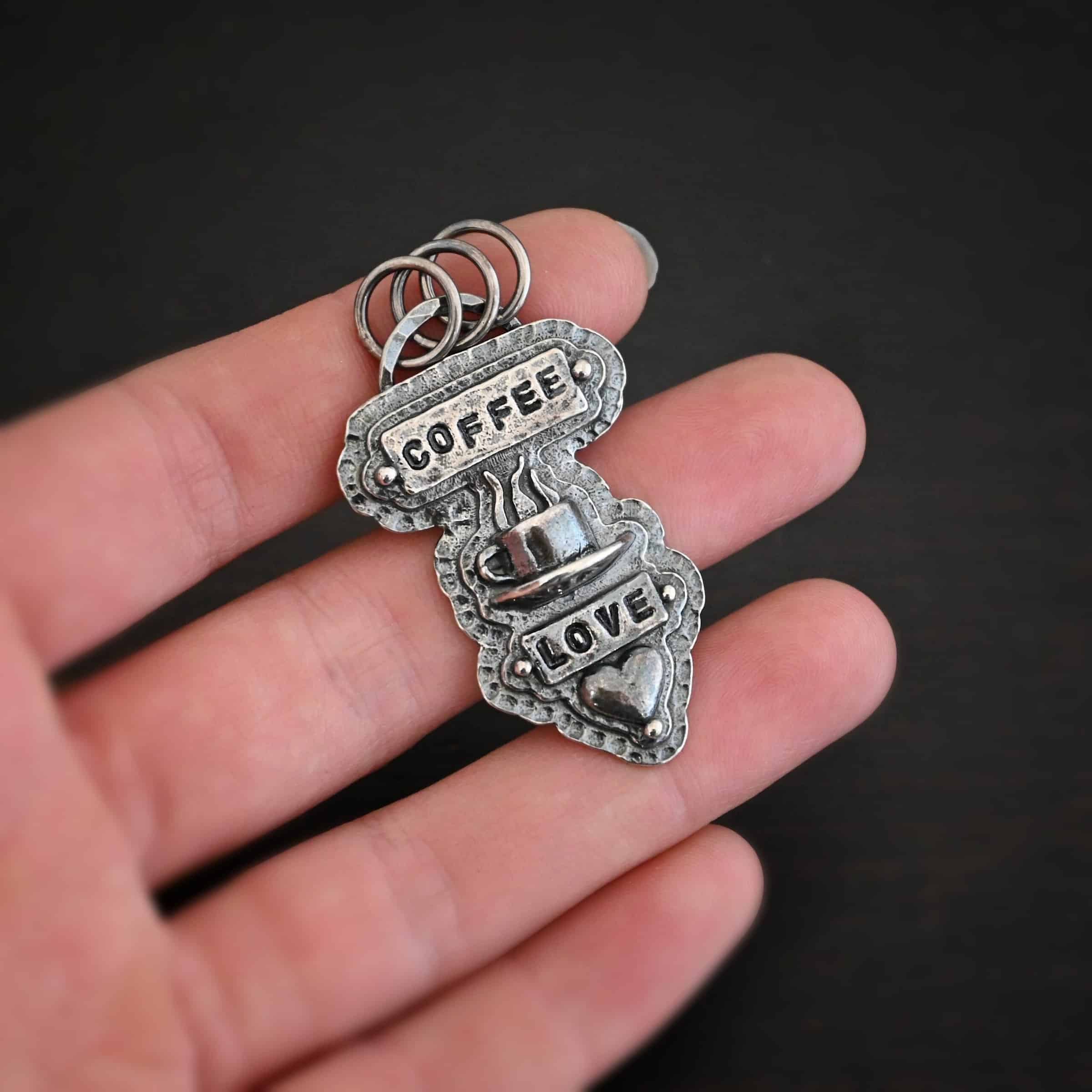 True Addiction Coffee Necklace Photo of the "True Addiction" Coffee Necklace. The pendant has two layers of sterling silver, both of which are hammered. The top layer is smaller than the bottom layer, so that both are noticeable. The top layer has a rectangle bar of silver soldered to it that has the word "Coffee" stamped into it. On each end of the bar is a sterling silver ball. Beneath the bar, soldered to the top layer is a coffee cup charm sitting on a saucer with steam waves coming out of the top of it. Beneath the saucer of the cup is another smaller rectangle bar on the top layer that has "love" stamped into it. There is a silver ball on each end of the rectangle. Beneath the rectangle is a heart charm that is soldered. Beneath the heart is a silver ball on the tip of the heart. The top of the necklace, on the back, has two loops, one loop on each side. Attached to each loop is the chain of the necklace. Necklace is lying against the fingertips of a hand to demonstrate size.