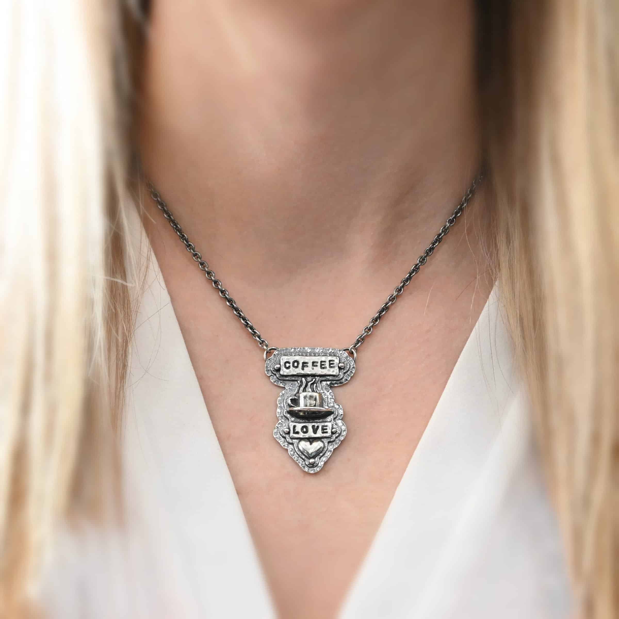 True Addiction Coffee Necklace Photo of the "True Addiction" Coffee Necklace. The pendant has two layers of sterling silver, both of which are hammered. The top layer is smaller than the bottom layer, so that both are noticeable. The top layer has a rectangle bar of silver soldered to it that has the word "Coffee" stamped into it. On each end of the bar is a sterling silver ball. Beneath the bar, soldered to the top layer is a coffee cup charm sitting on a saucer with steam waves coming out of the top of it. Beneath the saucer of the cup is another smaller rectangle bar on the top layer that has "love" stamped into it. There is a silver ball on each end of the rectangle. Beneath the rectangle is a heart charm that is soldered. Beneath the heart is a silver ball on the tip of the heart. The top of the necklace, on the back, has two loops, one loop on each side. Attached to each loop is the chain of the necklace. Necklace is worn by a model to demonstrate size.