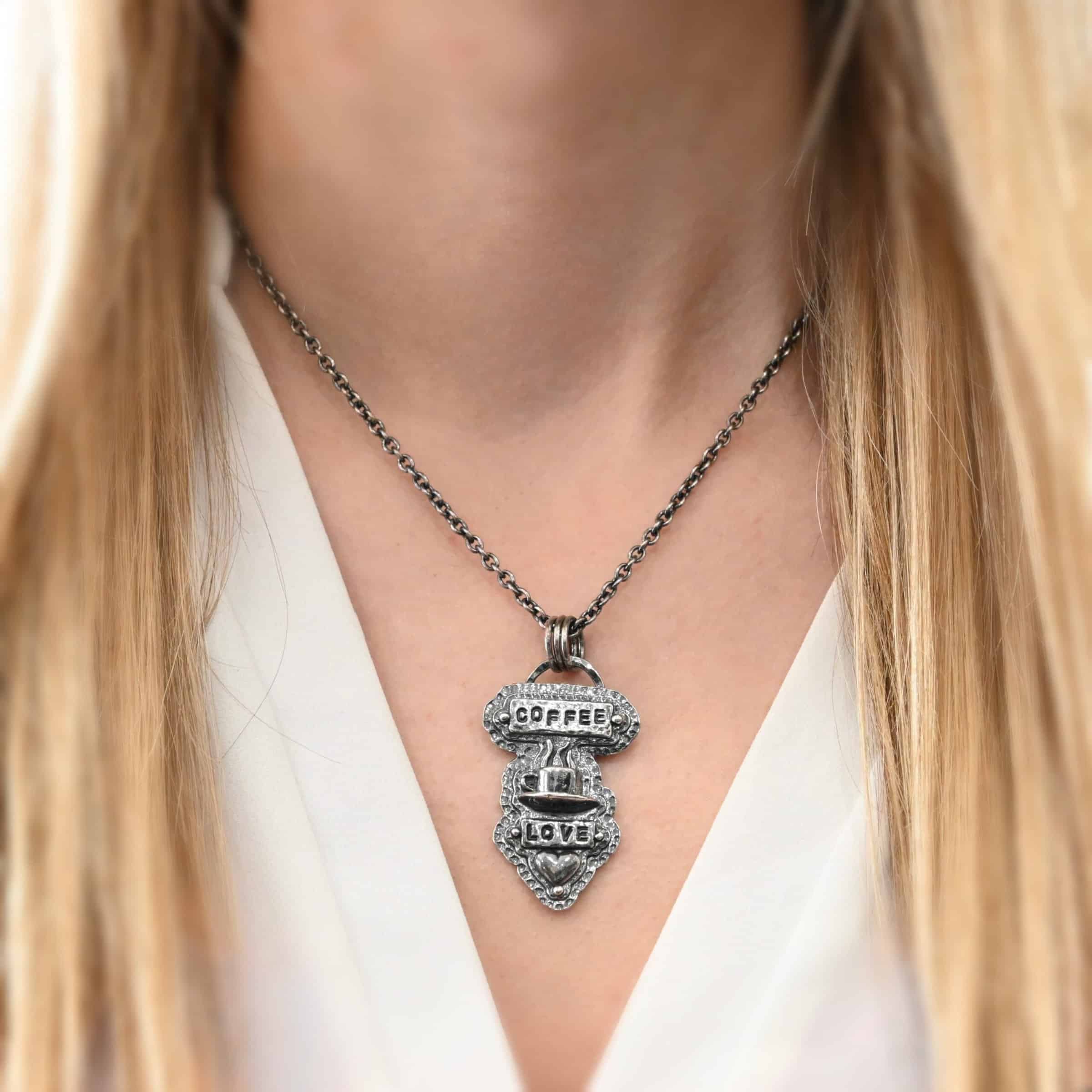 True Addiction Coffee Necklace Photo of the "True Addiction" Coffee Necklace. The pendant has two layers of sterling silver, both of which are hammered. The top layer is smaller than the bottom layer, so that both are noticeable. The top layer has a rectangle bar of silver soldered to it that has the word "Coffee" stamped into it. On each end of the bar is a sterling silver ball. Beneath the bar, soldered to the top layer is a coffee cup charm sitting on a saucer with steam waves coming out of the top of it. Beneath the saucer of the cup is another smaller rectangle bar on the top layer that has "love" stamped into it. There is a silver ball on each end of the rectangle. Beneath the rectangle is a heart charm that is soldered. Beneath the heart is a silver ball on the tip of the heart. The top of the necklace, on the back, has two loops, one loop on each side. Attached to each loop is the chain of the necklace. Necklace is worn by a model to demonstrate size.