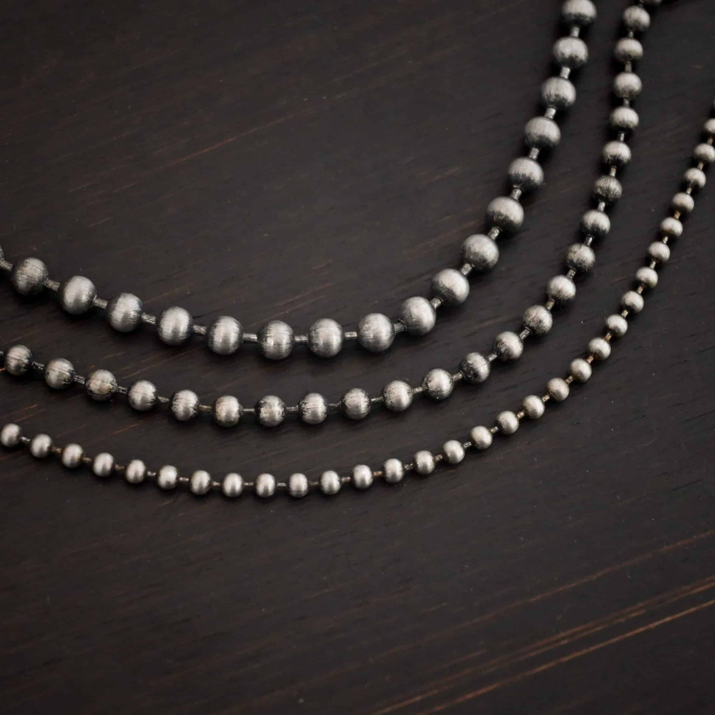 Beau Ball Chain Close up of the "Beau Ball" Chain with 4mm, 3.2 mm, and 2.4mm ball chains. Made out of brushed stainless steel with balls that are spaced apart evenly on the chain. Chains are shown against a dark background. The highest chain in the photo is the 4mm chain, with the 3.2mm chain in the middle, and the 2.4mm chain at the bottom of the photo.
