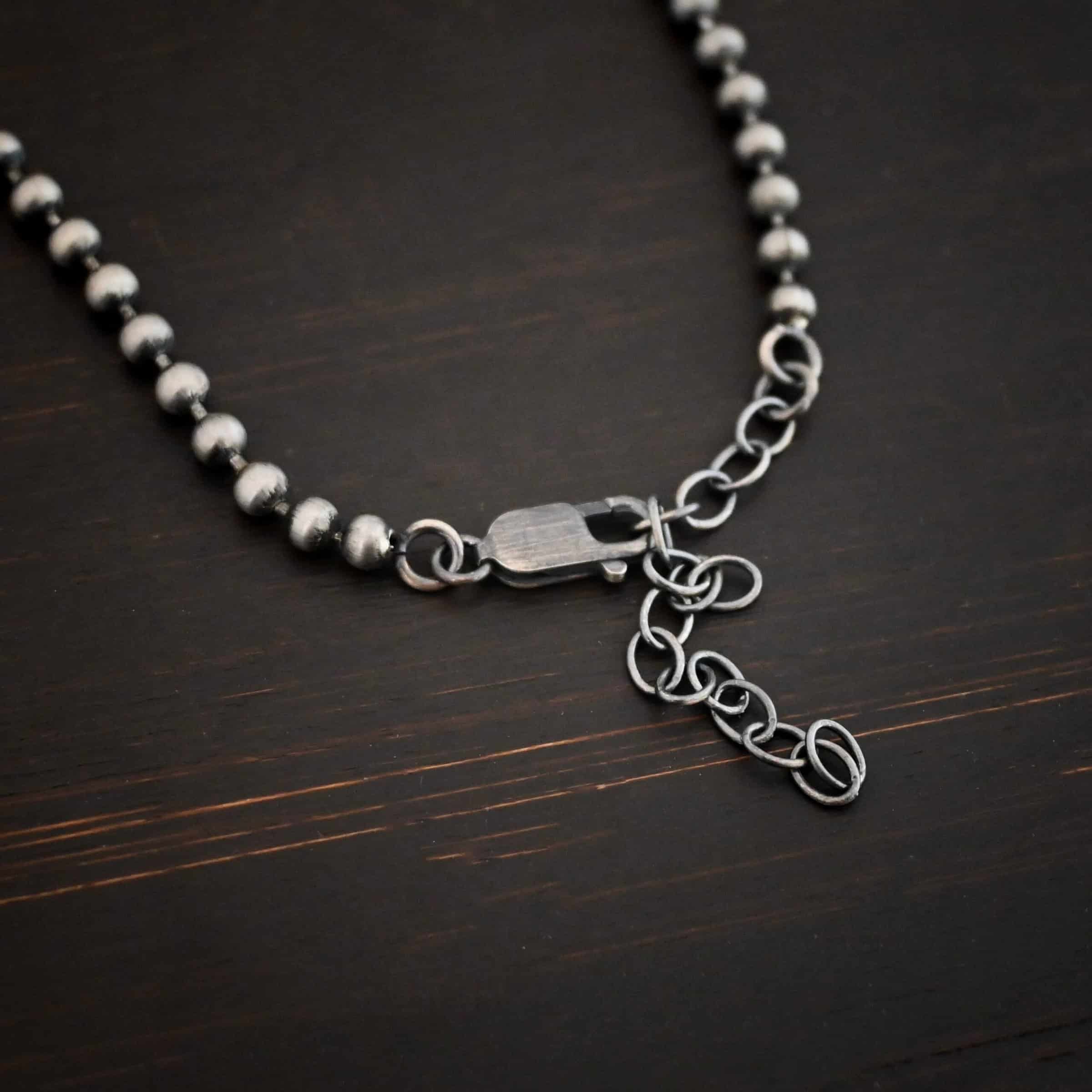 Beau Ball Chain Close up of the "Beau Ball" Chain. Made out of brushed stainless steel with 4mm balls that are spaced apart evenly on the chain. Chain is set against a dark background. The lobster claw clasp is the main part of the photo with the extension for the chain.