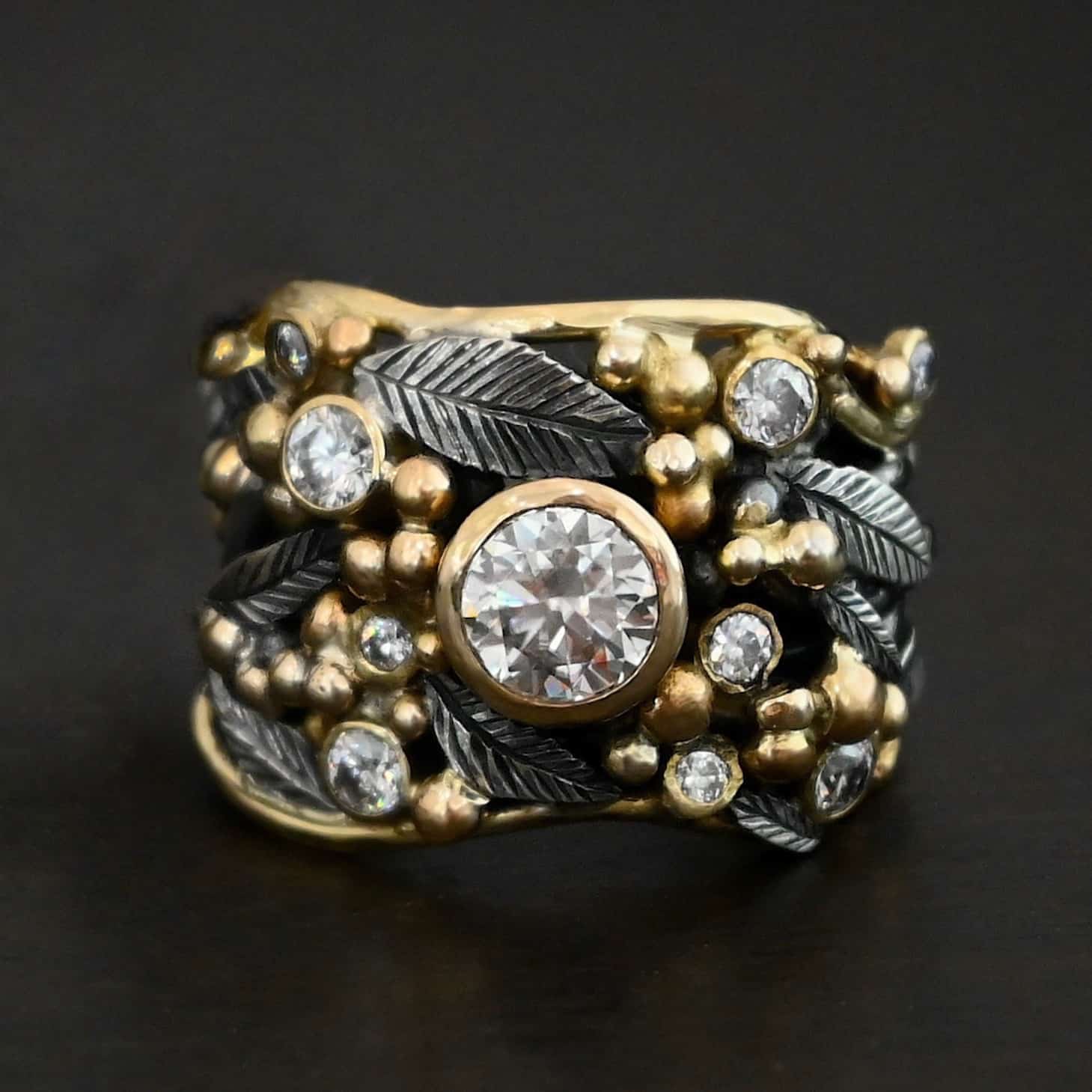 Photo of the "Enchanted" Ring. Ring has a large moissanite in the center and smaller moissanites scattered around the entire ring. There are multiple silver leaves around the entire ring, and a ton of gold ball detail around the ring. Each of the stones are set in gold. There are seven stones total, the center stone, and six moissanites around the rest of the ring, three on each side. Ring is set against a dark background.