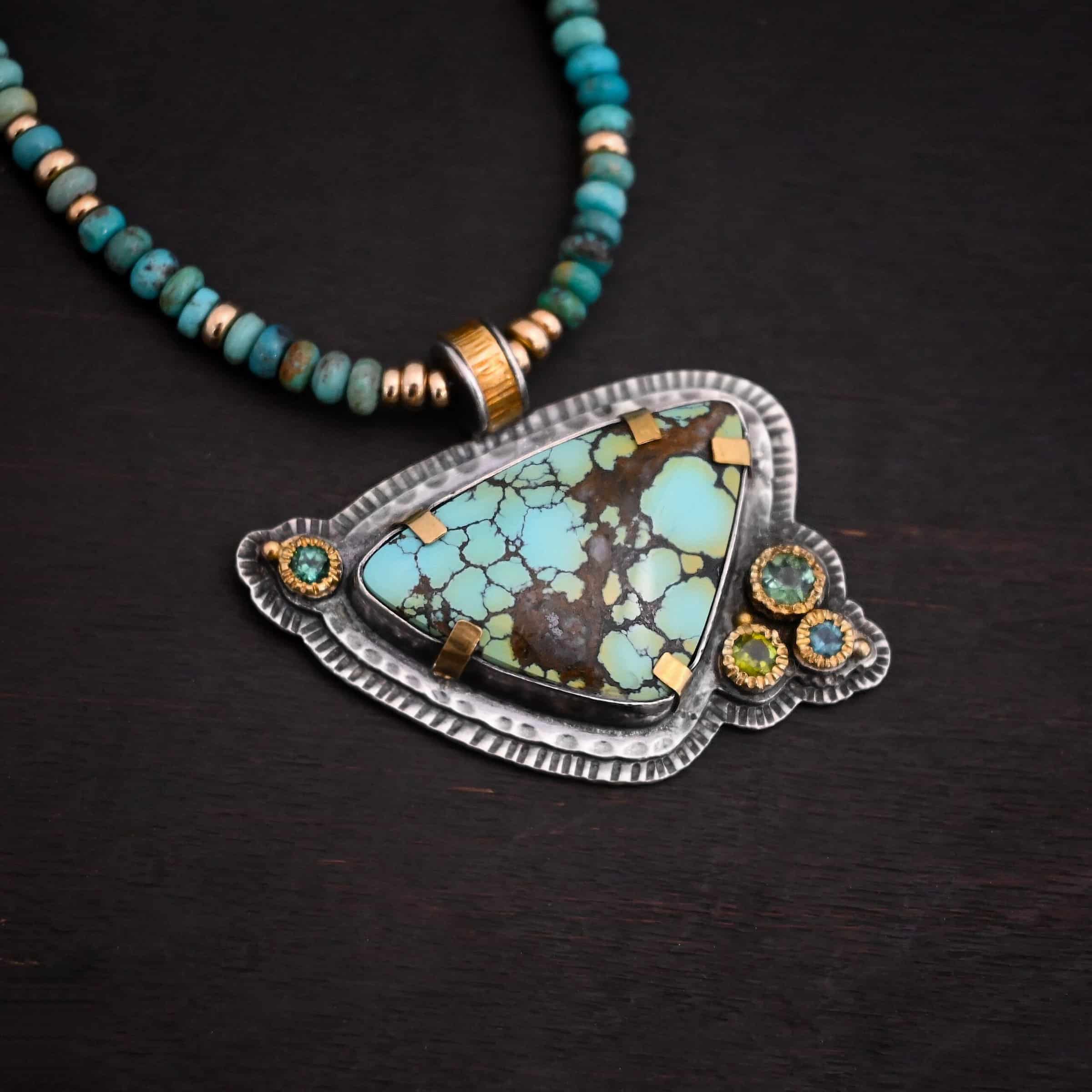 Close up of the "Trinity" Necklace. Made out of sterling silver, gold, turquoise, and tourmaline. The turquoise is the center stone. It is a mix of blue dots with brown and green scattered throughout the stone. The turquoise has gold tabs around it to help keep it held into place. There are four tourmalines that are a mix of blue and green colors. They are all set in gold. Three of them are on the bottom right of the necklace, and one is in the top left corner of the necklace. There are two layers of hammered sterling silver that the turquoise and tourmalines are set on. The pendant has a medium sized hoop on the top of it that is gold, and is attached to the chain which is made out of turquoise and gold beads. Pendant is laying against a dark background.