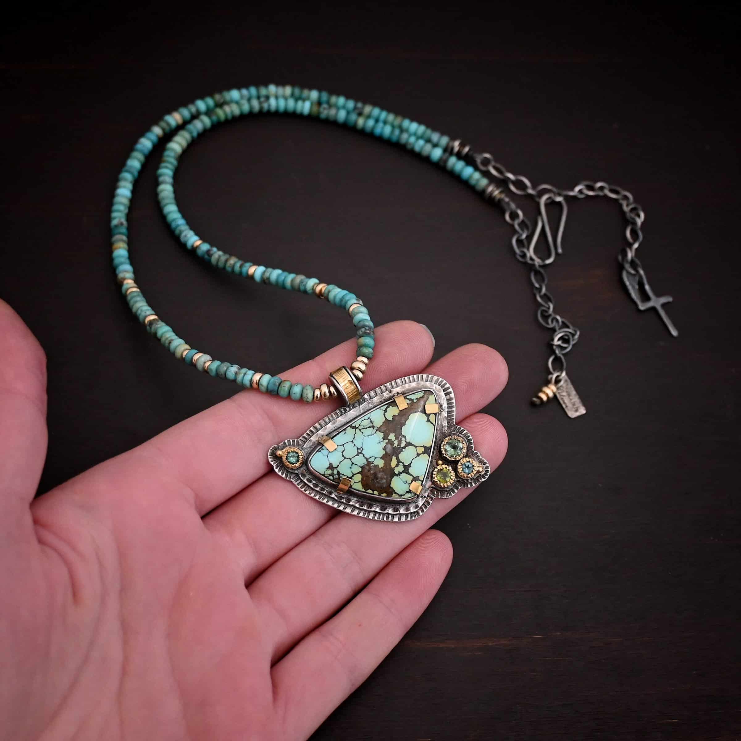 Close up of the "Trinity" Necklace. Made out of sterling silver, gold, turquoise, and tourmaline. The turquoise is the center stone. It is a mix of blue dots with brown and green scattered throughout the stone. The turquoise has gold tabs around it to help keep it held into place. There are four tourmalines that are a mix of blue and green colors. They are all set in gold. Three of them are on the bottom right of the necklace, and one is in the top left corner of the necklace. There are two layers of hammered sterling silver that the turquoise and tourmalines are set on. The pendant has a medium sized hoop on the top of it that is gold, and is attached to the chain which is made out of turquoise and gold beads. Pendant is laying against the fingertips of a hand.