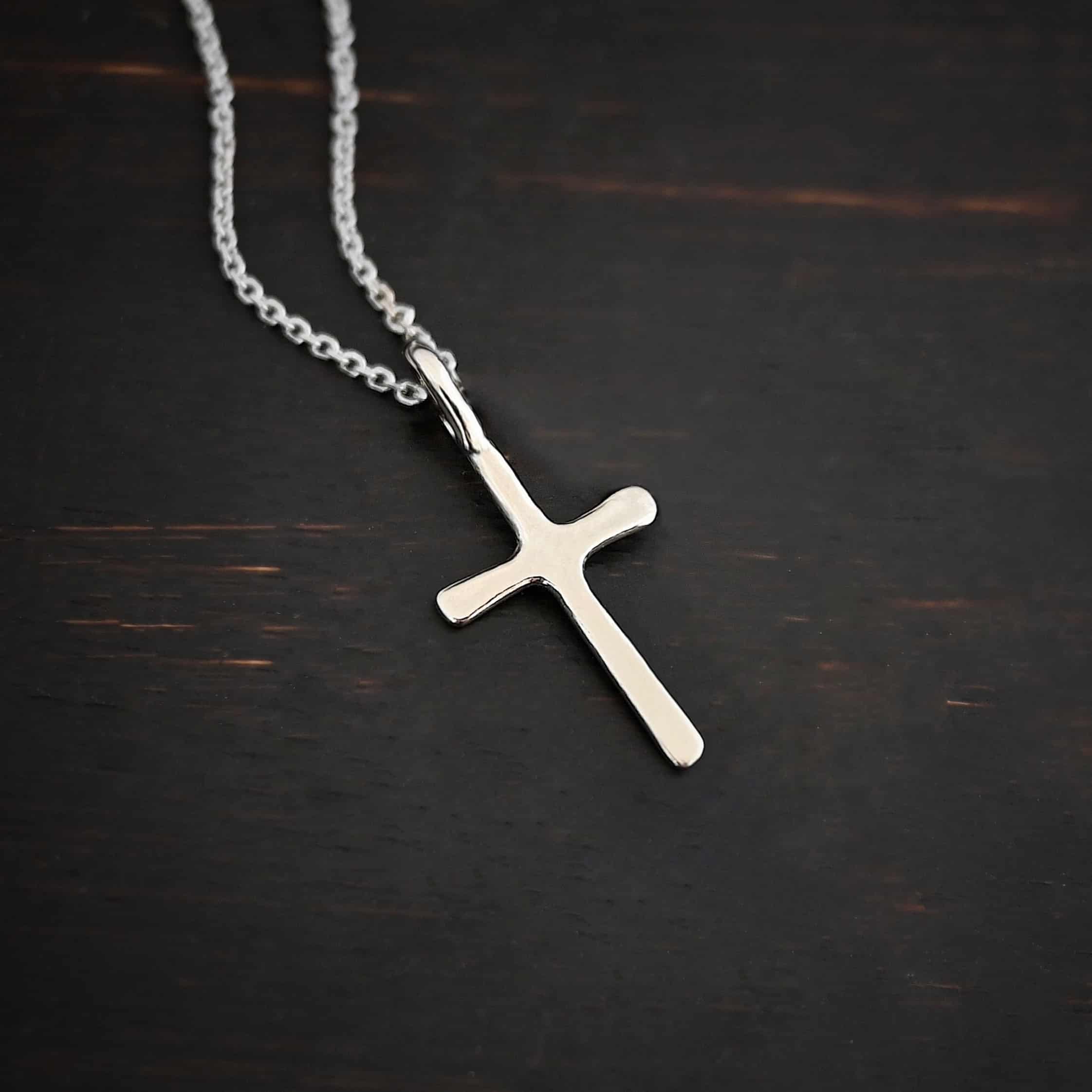 Divine Necklace Photo of the "Divine" Necklace. Made out of sterling silver. There is a small sized cross that is flat and smooth. It has a loop soldered to the top of it. There is a chain that goes through the loop of the cross that is also silver. Necklace is lying against a dark background.