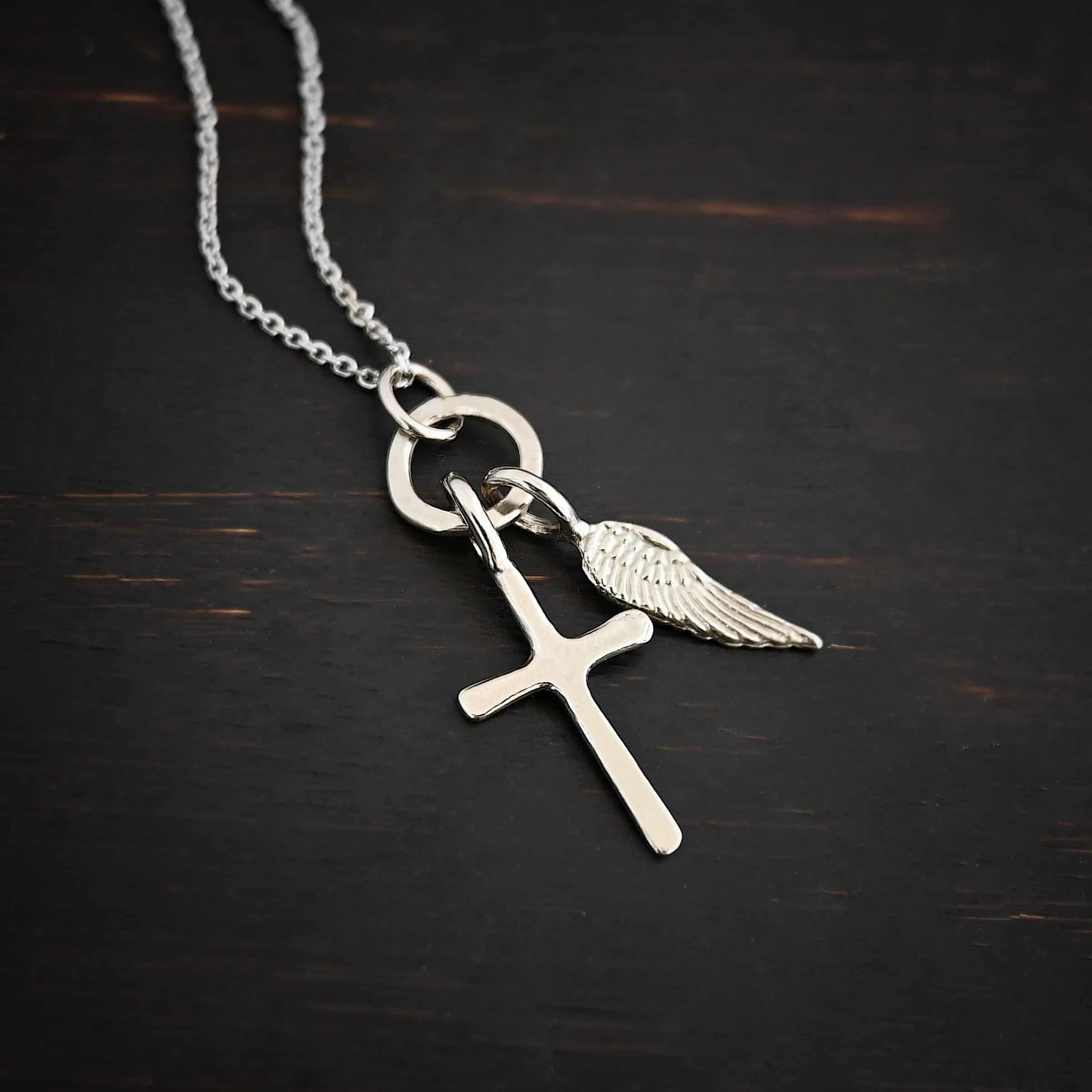 Divine Necklace Photo of the "Divine" Necklace. Made out of sterling silver. There is a small sized cross that is flat and smooth. It has a loop soldered to the top of it. There is a small sized angel wing pendant next to it, also with a soldered loop on the top. There is a chain that goes through the top of the necklace that is also silver. Necklace is lying against a dark background.