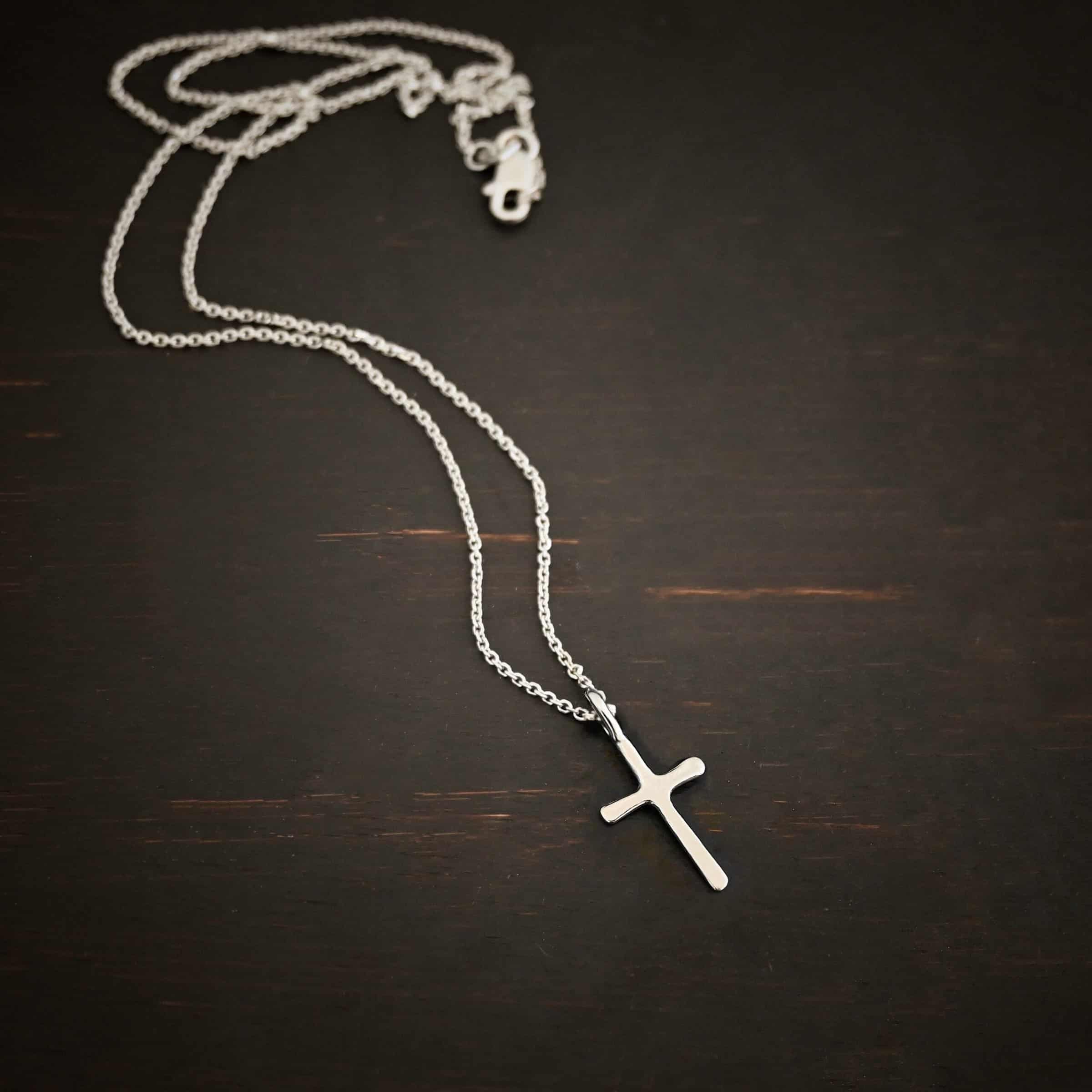 Divine Necklace Photo of the "Divine" Necklace. Made out of sterling silver. There is a small sized cross that is flat and smooth. It has a loop soldered to the top of it. There is a chain that goes through the loop of the cross that is also silver. Necklace is lying against a dark background.