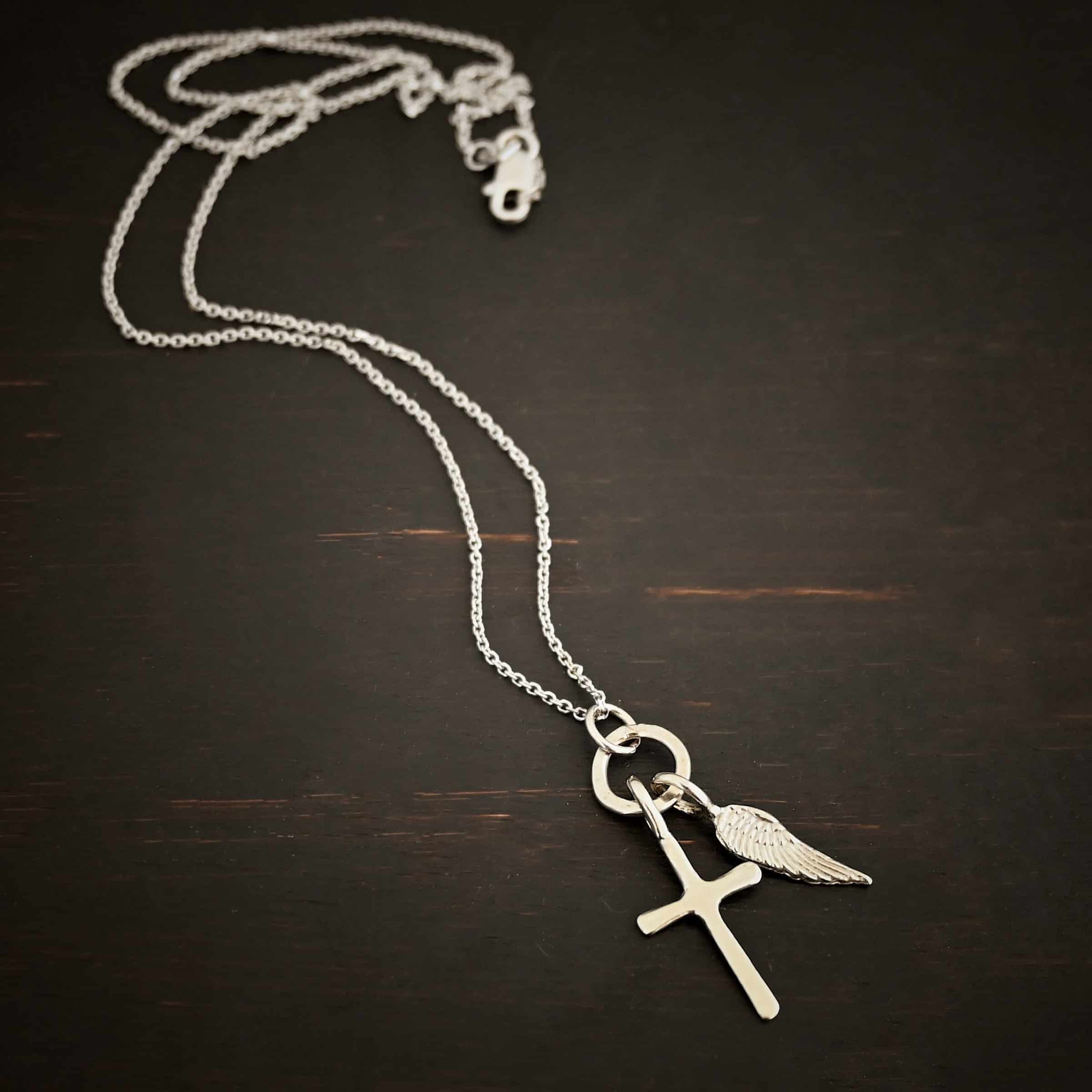 Divine Necklace Photo of the "Divine" Necklace. Made out of sterling silver. There is a small sized cross that is flat and smooth. It has a loop soldered to the top of it. There is a small sized angel wing pendant next to it, also with a soldered loop on the top. There is a chain that goes through the top of the necklace that is also silver. Necklace is lying against a dark background.