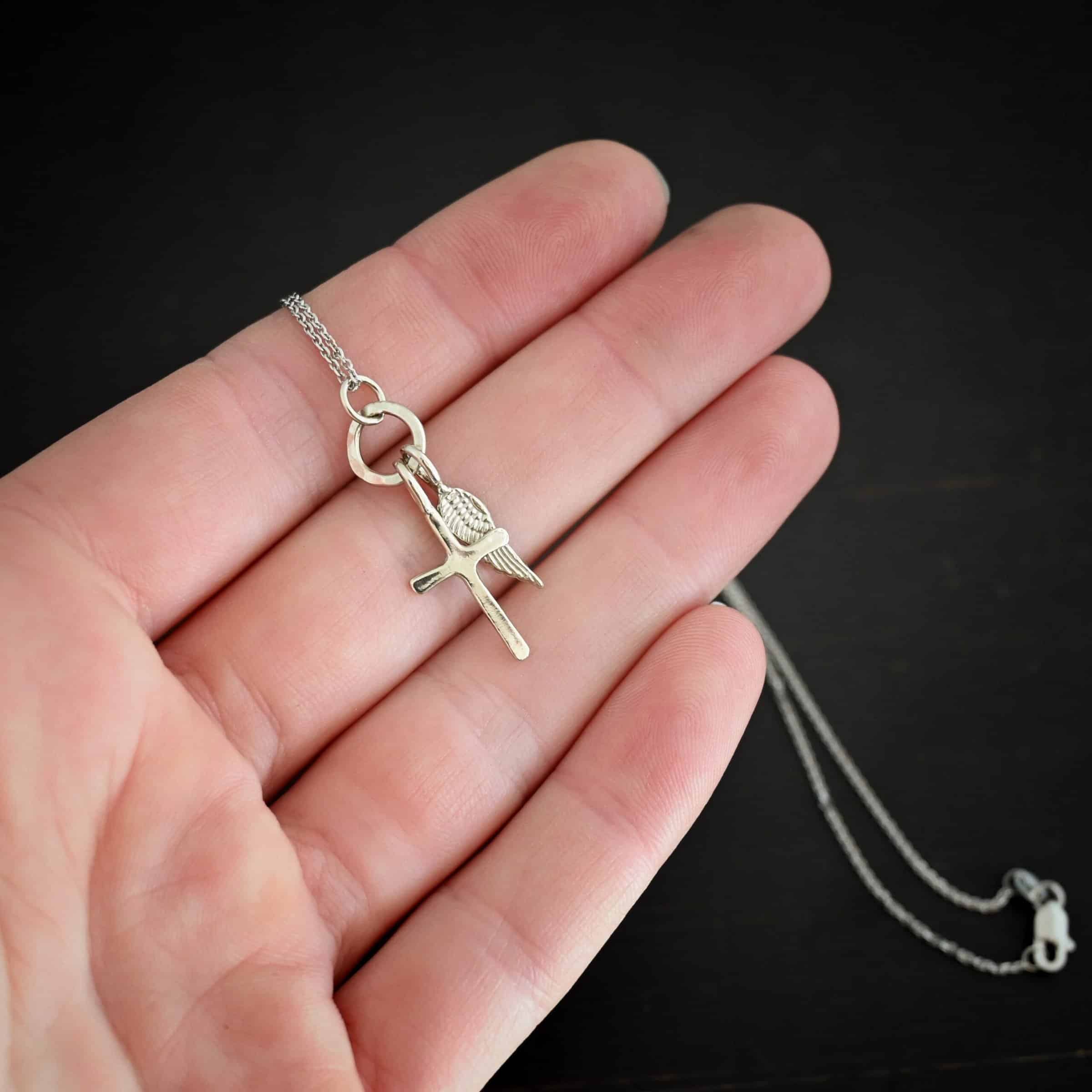 Divine Necklace Photo of the "Divine" Necklace. Made out of sterling silver. There is a small sized cross that is flat and smooth. It has a loop soldered to the top of it. There is a small sized angel wing pendant next to it, also with a soldered loop on the top. There is a chain that goes through the top of the necklace that is also silver. Necklace is lying against the fingertips of a hand.