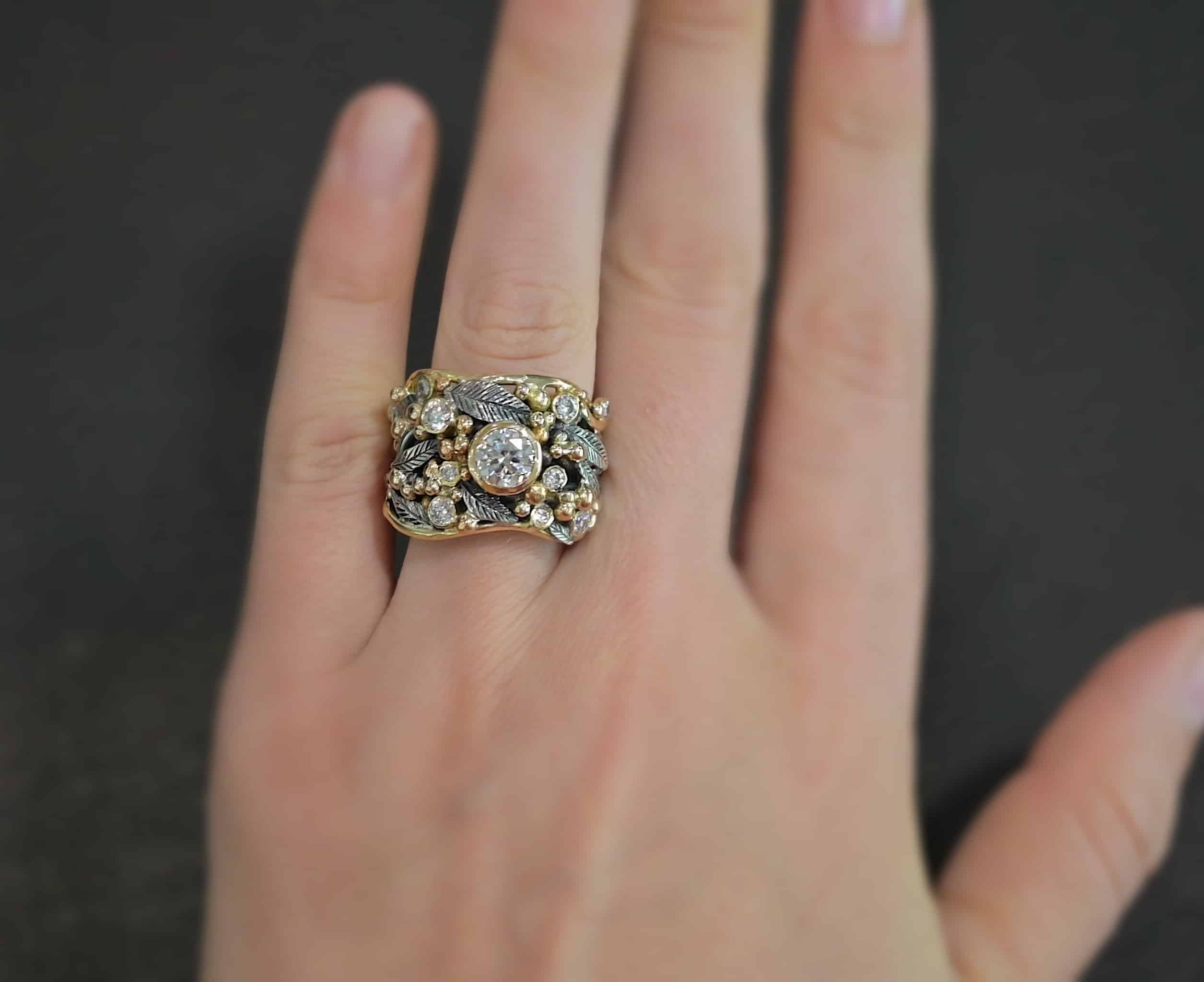 Photo of the "Enchanted" Ring. Ring has a large moissanite in the center and smaller moissanites scattered around the entire ring. There are multiple silver leaves around the entire ring, and a ton of gold ball detail around the ring. Each of the stones are set in gold. There are seven stones total, the center stone, and six moissanites around the rest of the ring, three on each side. Ring is worn on a models engagement ring finger to demonstrate size.